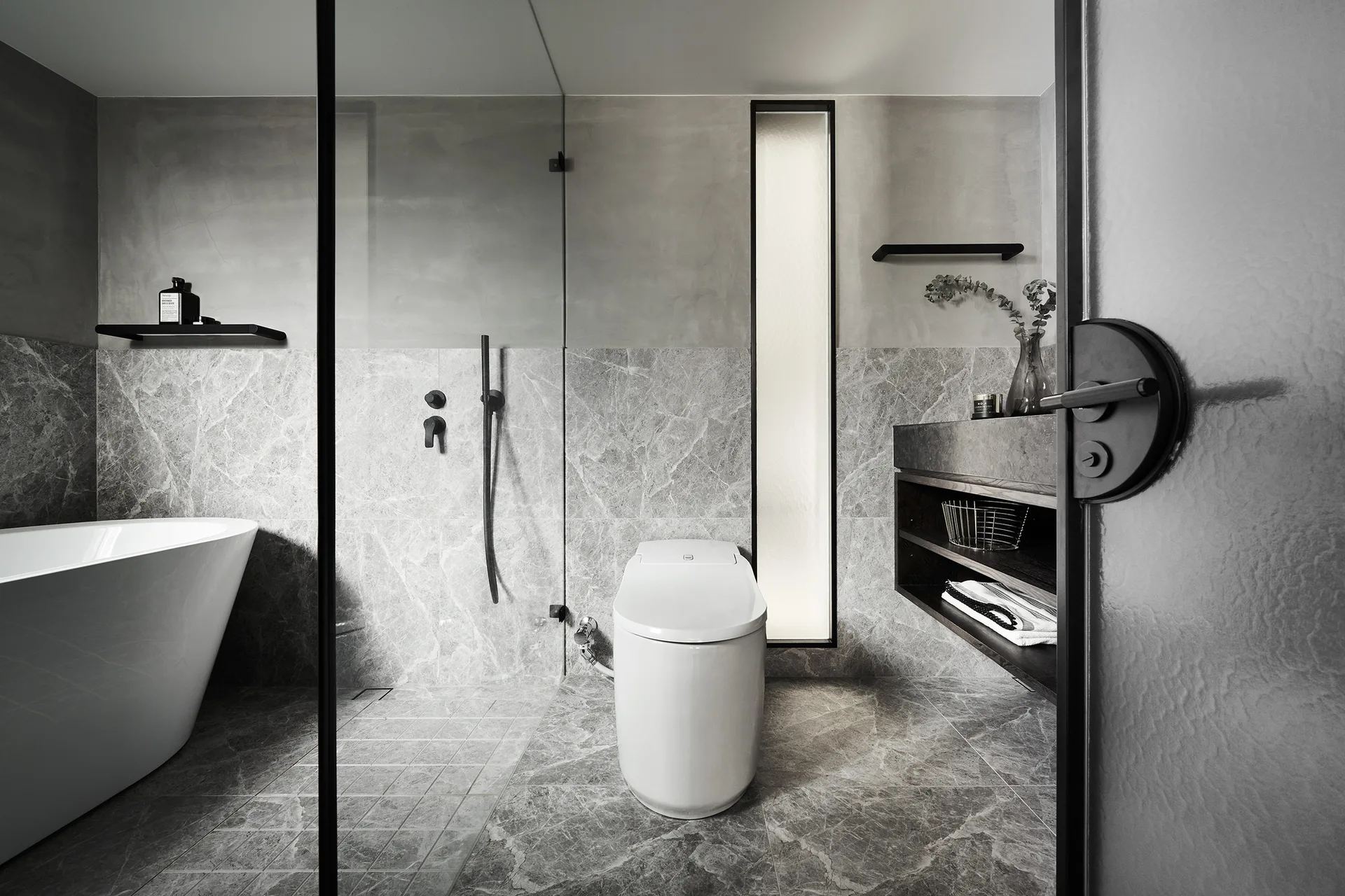A modern bathroom featuring grey marble walls and floors, a freestanding white bathtub, a toilet, and a dark wood floating vanity.