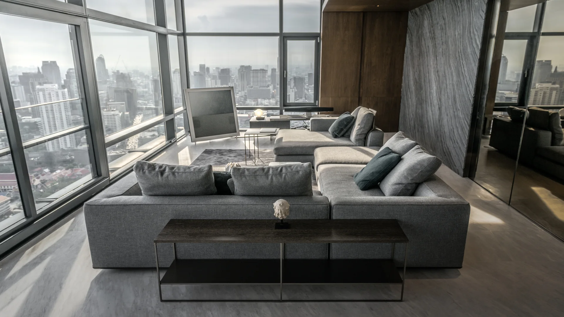 A minimalist living room features a large grey modular sectional sofa facing floor-to-ceiling windows with a city skyline view.
