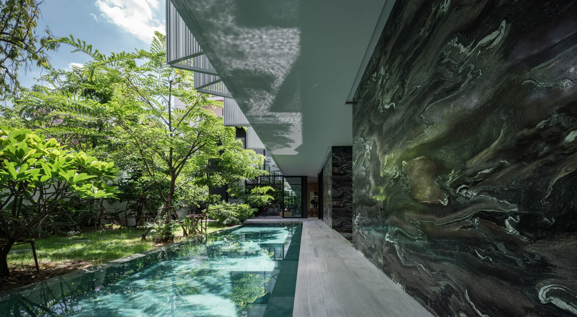 A long rectangular swimming pool runs parallel to a large green veined stone feature wall and a grey stone walkway under a deep overhang.