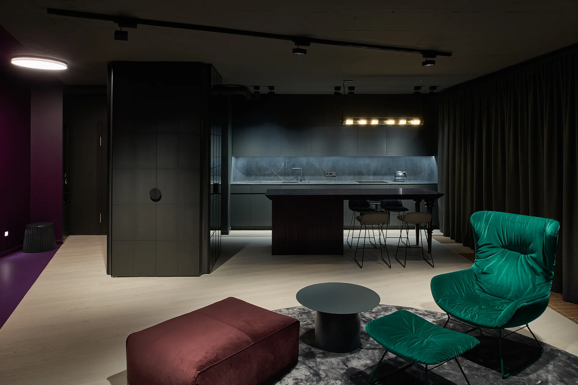 A modern open-plan room featuring a matte black kitchen and a living area with an emerald green armchair and a burgundy velvet ottoman on light floors.