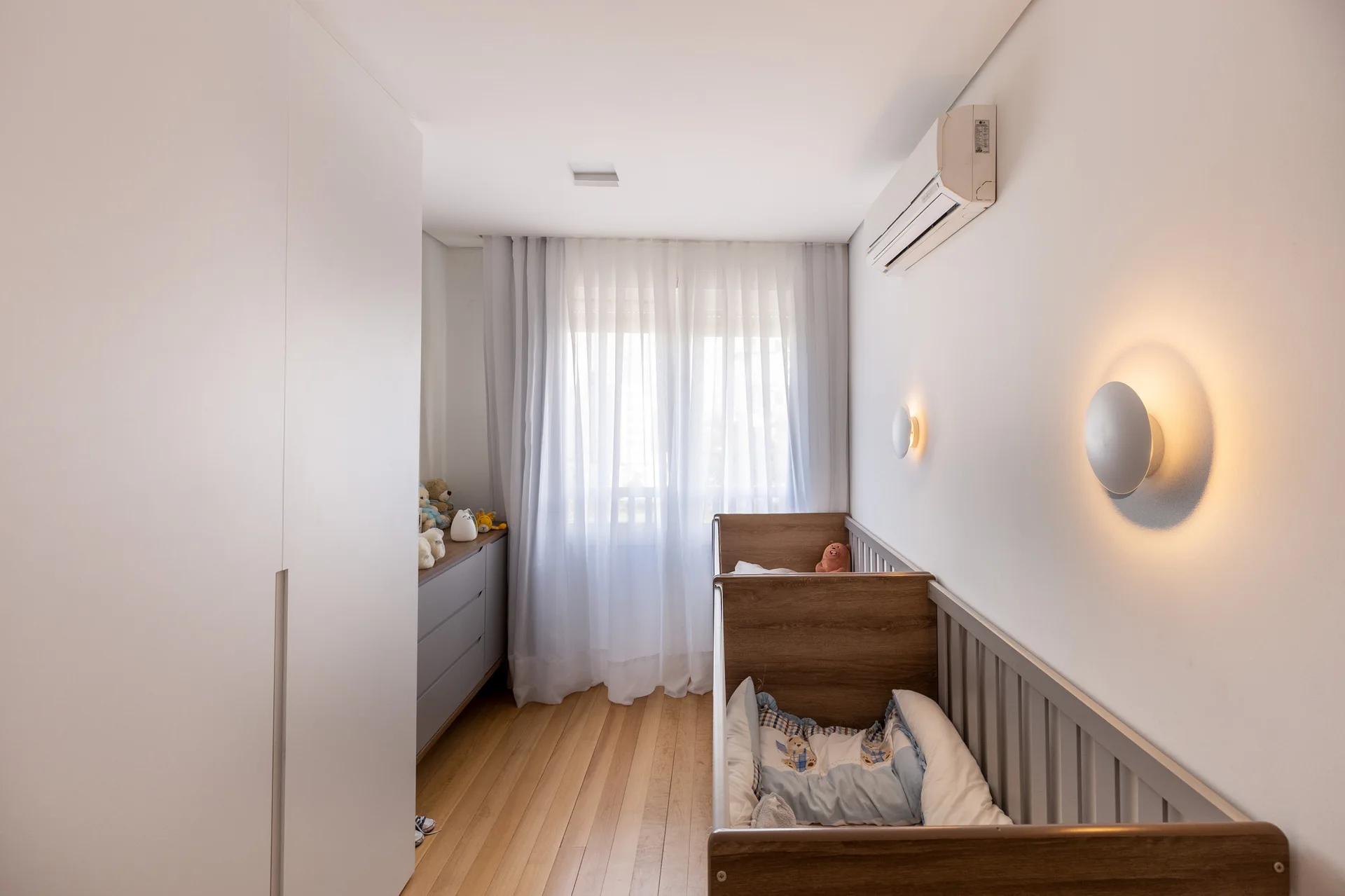 A modern minimalist nursery featuring a wood and grey crib, light wood flooring, warm circular wall sconces, and a white built-in wardrobe.