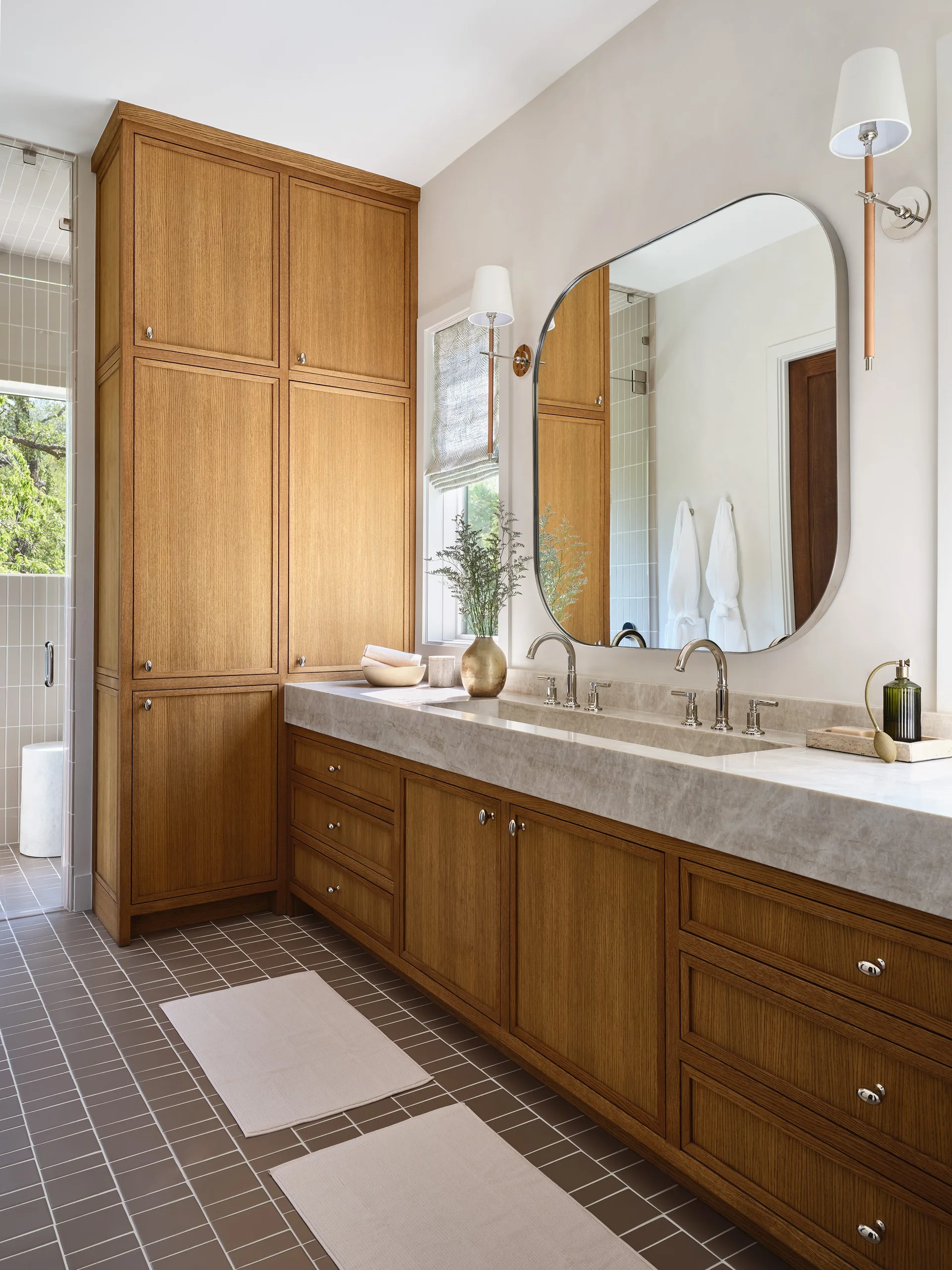 A contemporary bathroom features a wood double vanity with a stone countertop, a rounded silver-framed mirror, and a tall built-in oak storage cabinet.