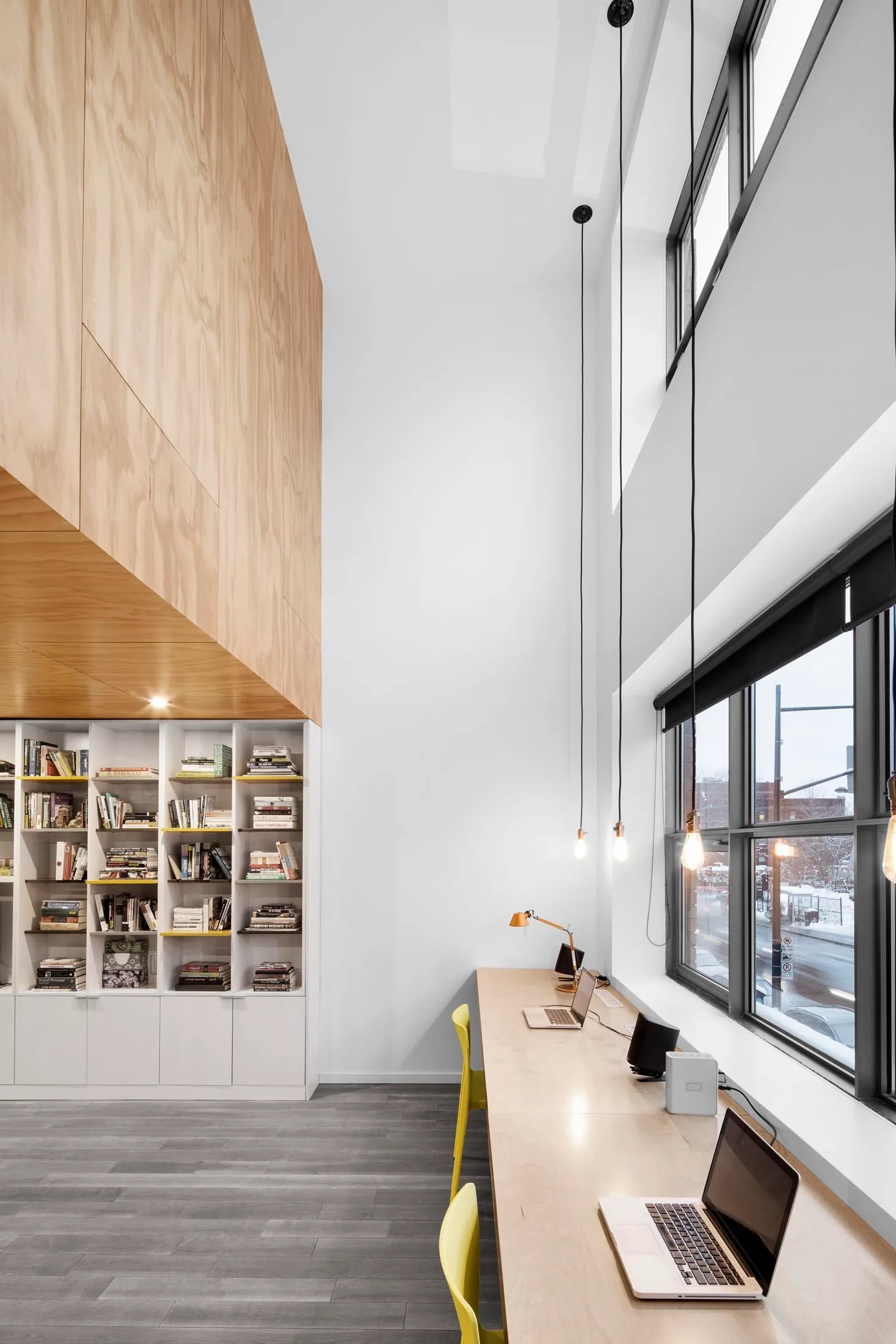 A high ceiling home office features a long light wood desk with yellow chairs, tall white bookshelves, and overhead wood wall paneling.