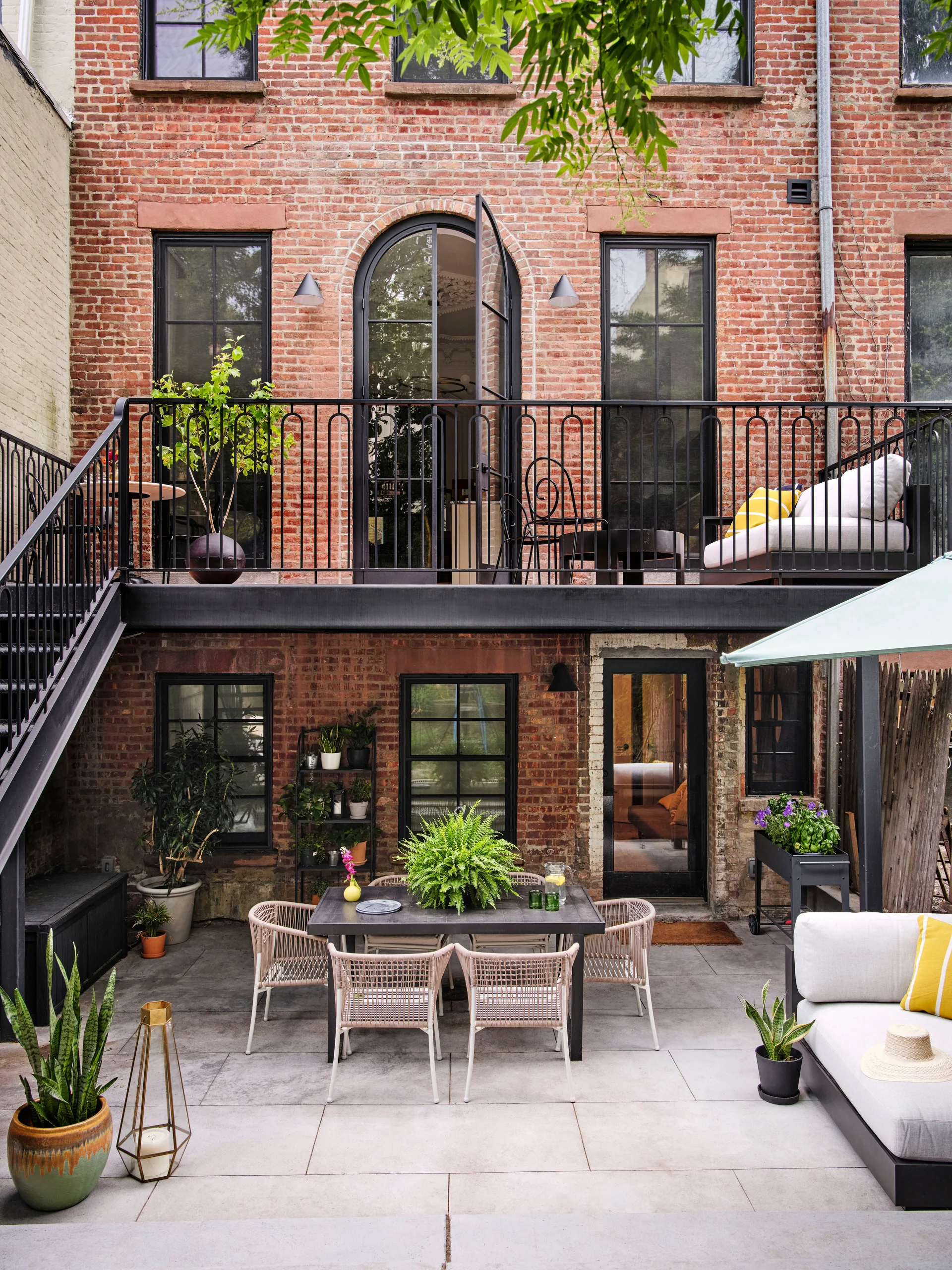 A paved urban patio features a black dining table, woven chairs, and a sectional sofa against a red brick townhouse with a black metal balcony.