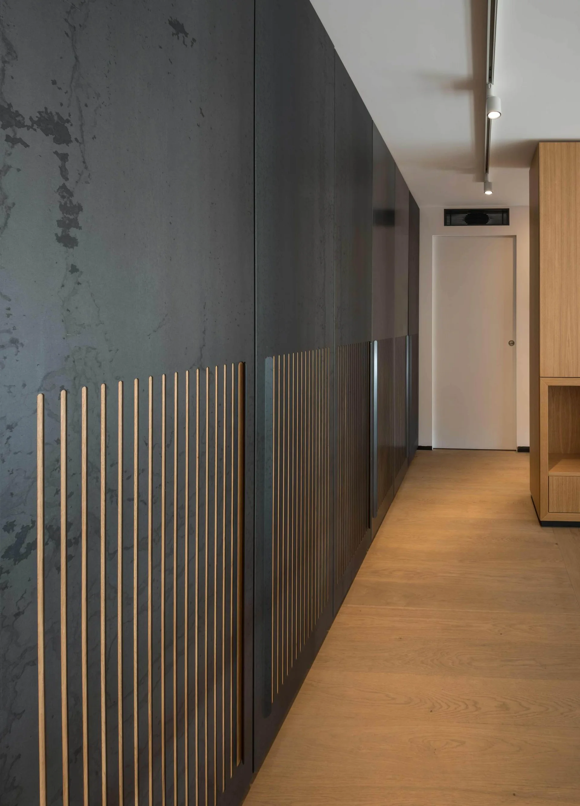 Dark grey floor-to-ceiling cabinets with vertical wood slat details line a hallway with light oak flooring and track lighting.