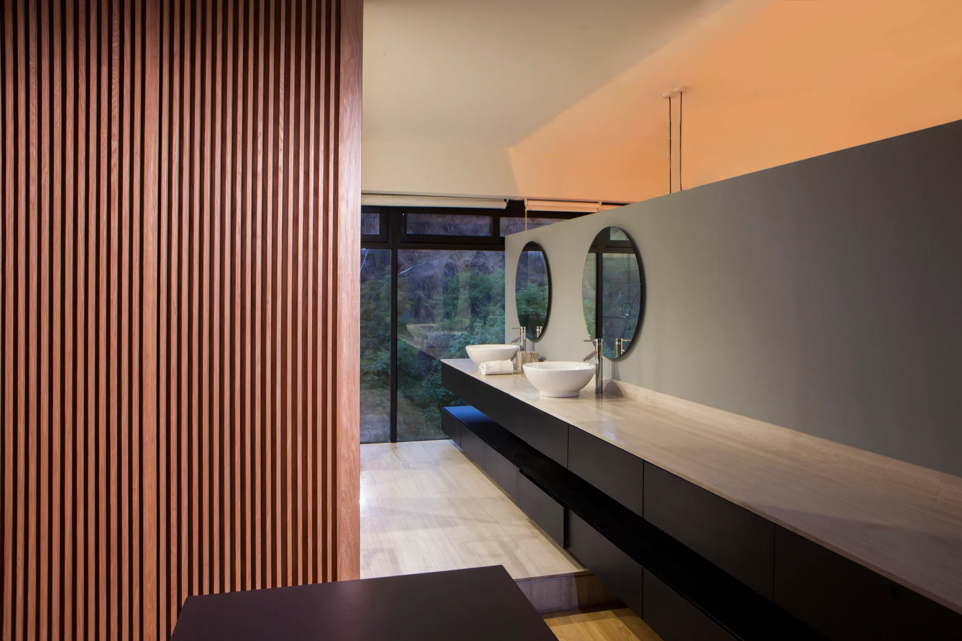 A modern bathroom features a long black floating vanity with two white vessel sinks and round mirrors next to a vertical wood slat partition.