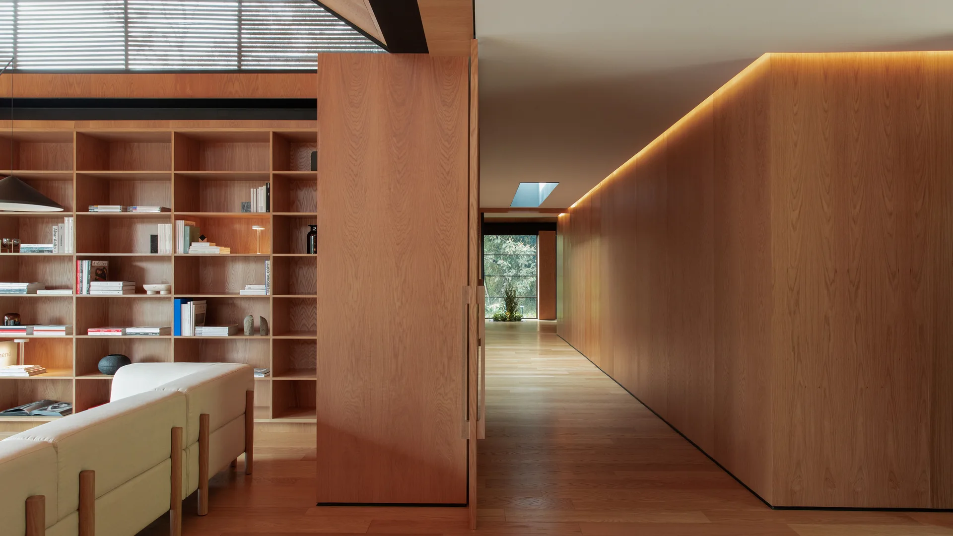 A modern living area featuring a light-colored sofa and wooden built-in bookshelf next to a long hallway lined with warm wood paneling.