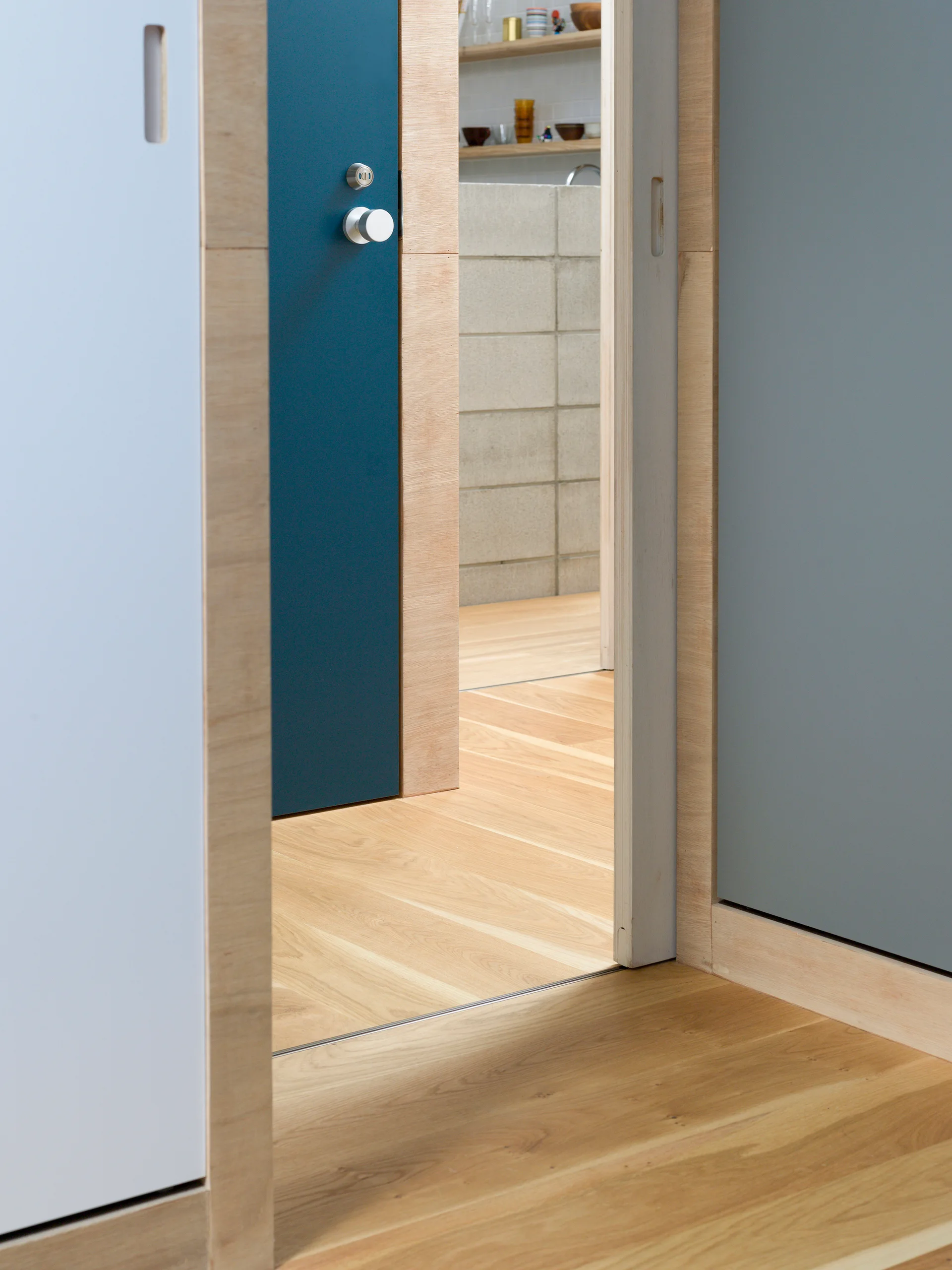 A view through a plywood-cased doorway with a deep teal door reveals a modern kitchen with a grey concrete block wall and natural wood flooring.