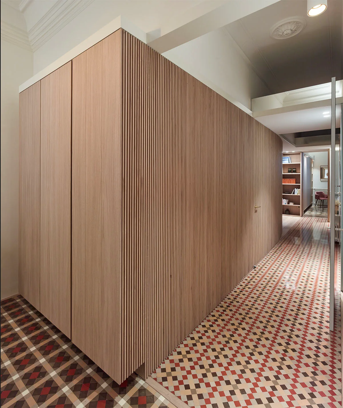 A long hallway features a floor-to-ceiling slatted wood cabinet block opposite white walls over a geometric red and grey tiled floor.
