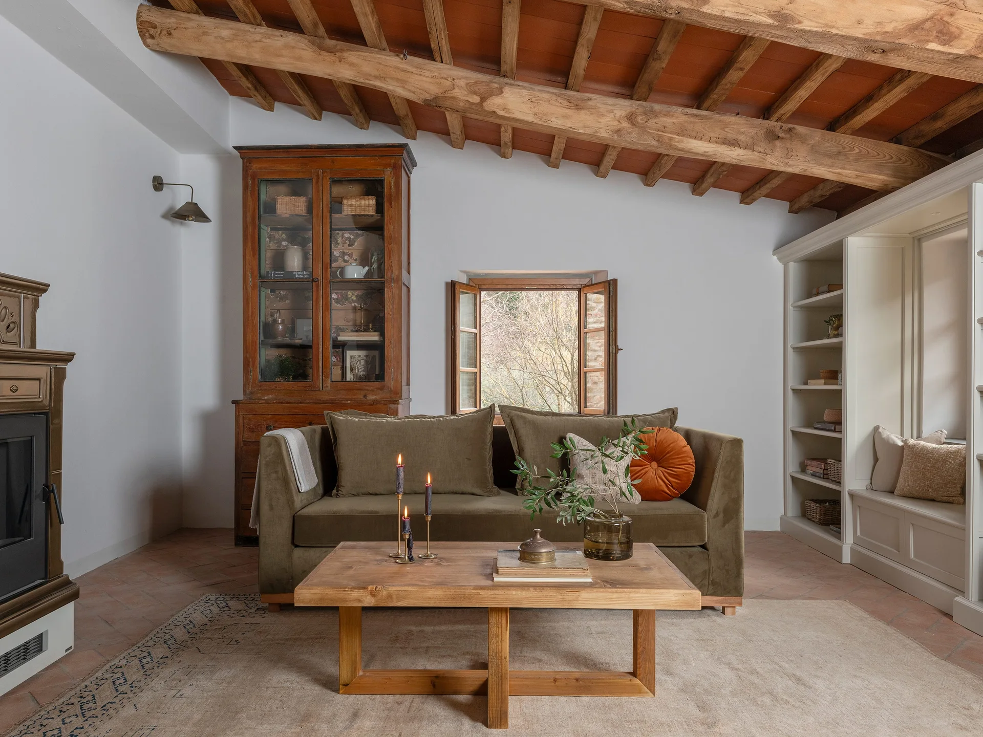 A rustic living room featuring a deep olive green sofa, a light wooden coffee table, terracotta floor tiles, and exposed ceiling beams.