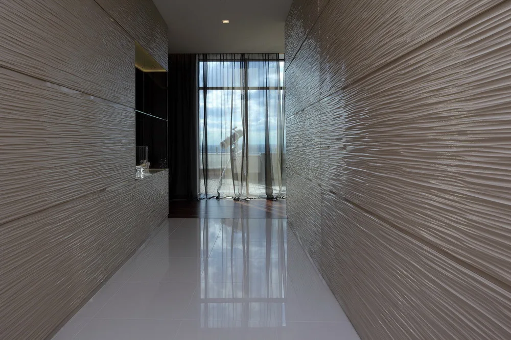 A modern hallway features wavy textured wall panels and a glossy reflective tile floor leading to a window with sheer curtains.