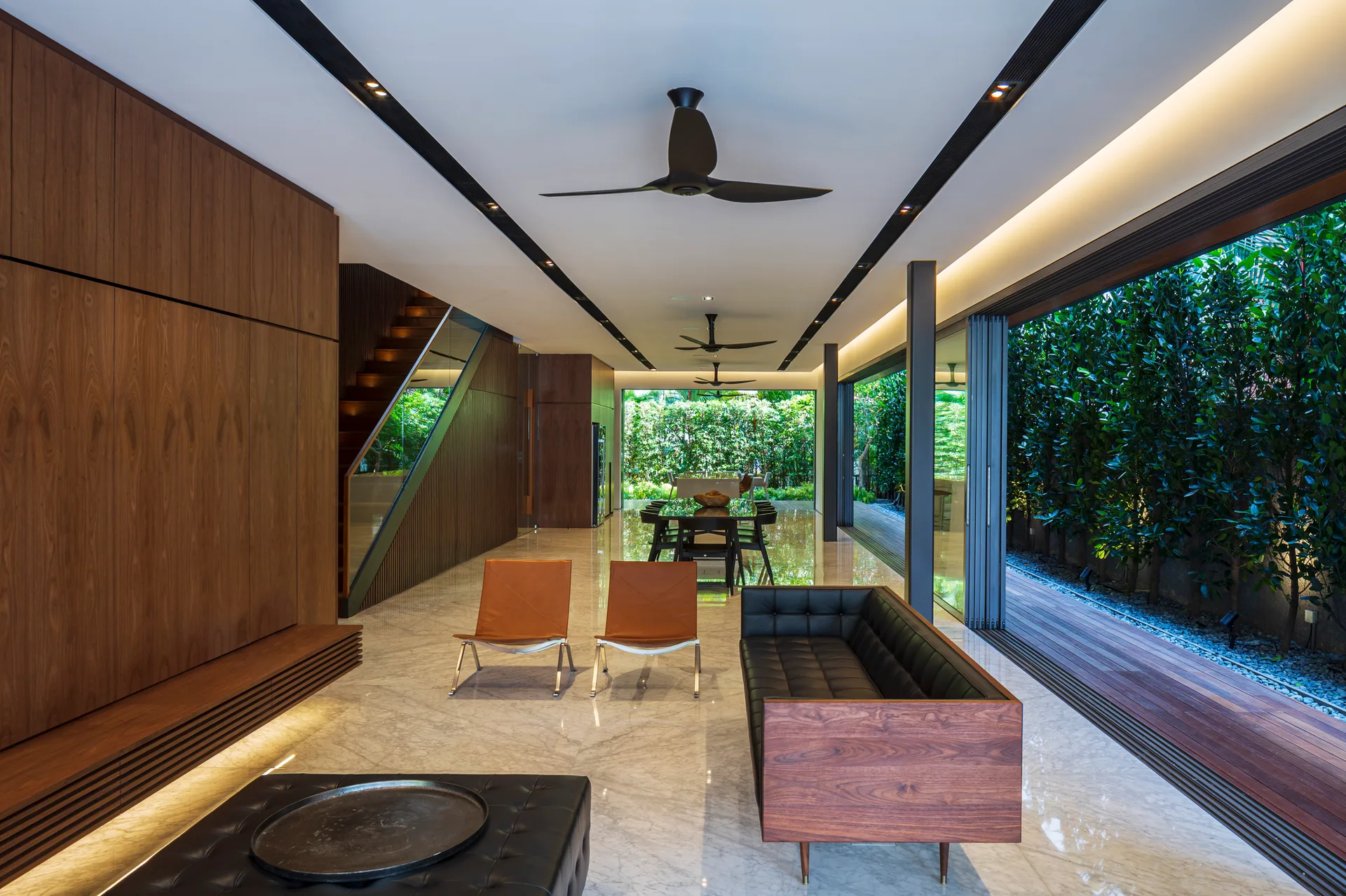 A modern open-plan living room featuring a wood-paneled wall, a black leather sofa, polished marble floors, and sliding glass doors opening to a garden.