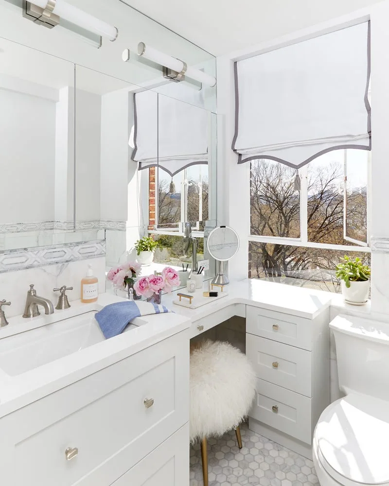 A bright white bathroom featuring a marble-topped L-shaped vanity, a fluffy white stool, and a window with a grey-bordered roman shade.