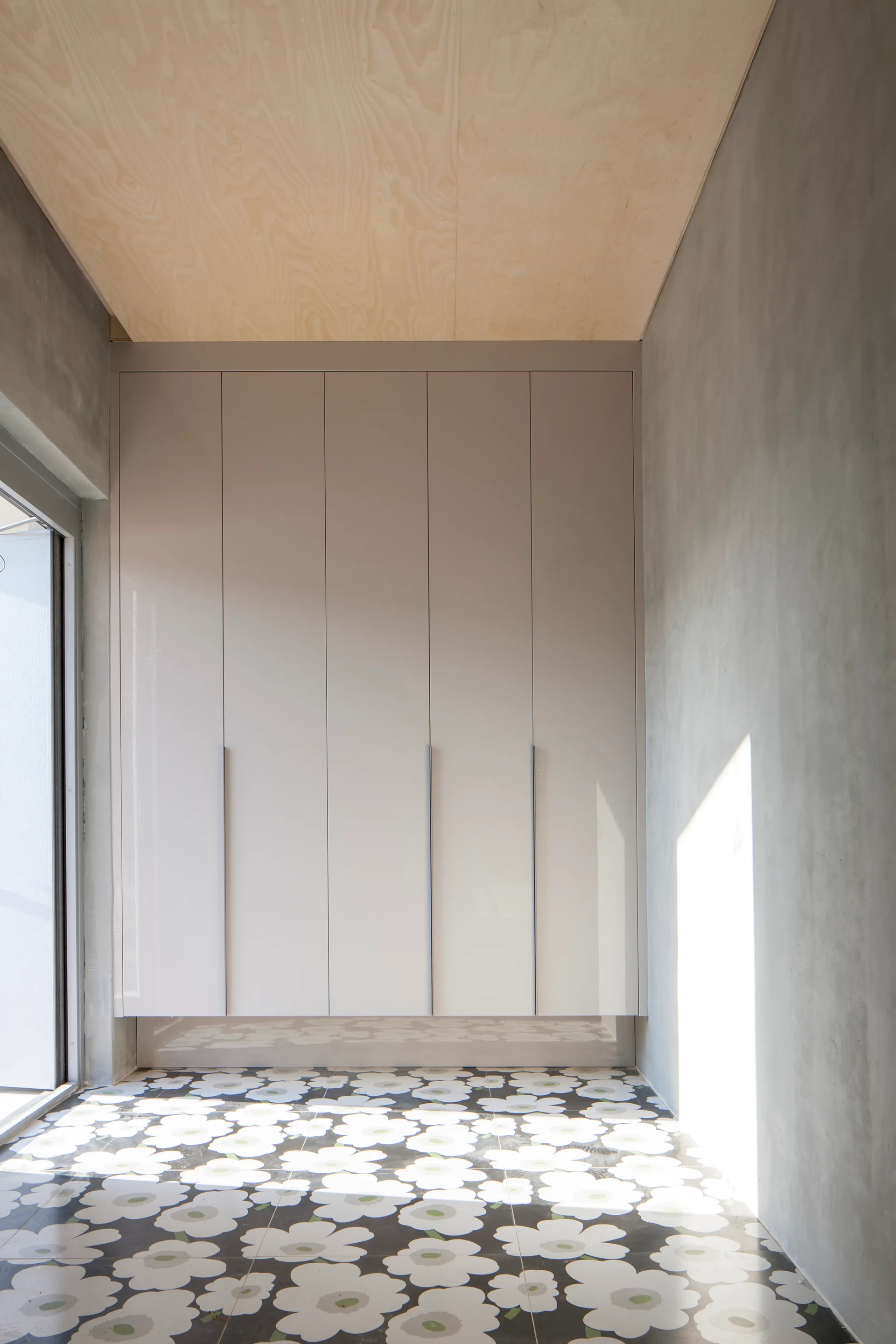 A minimalist hallway features a suspended off-white closet, textured grey concrete walls, a plywood ceiling, and black-and-white floral floor tiles.
