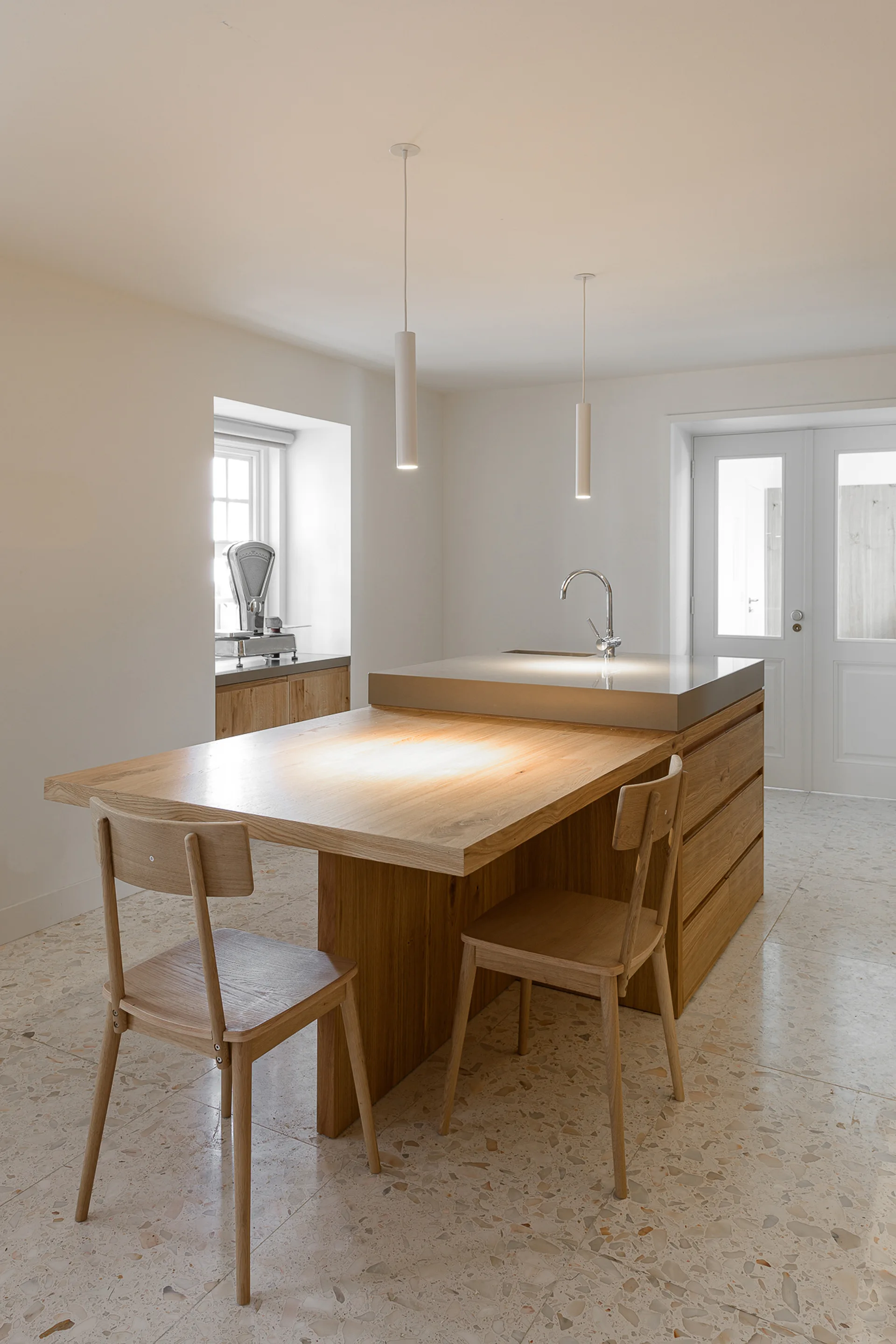 A modern Scandinavian kitchen with a dual-level oak and stone island, wooden dining chairs, and a light terrazzo floor.