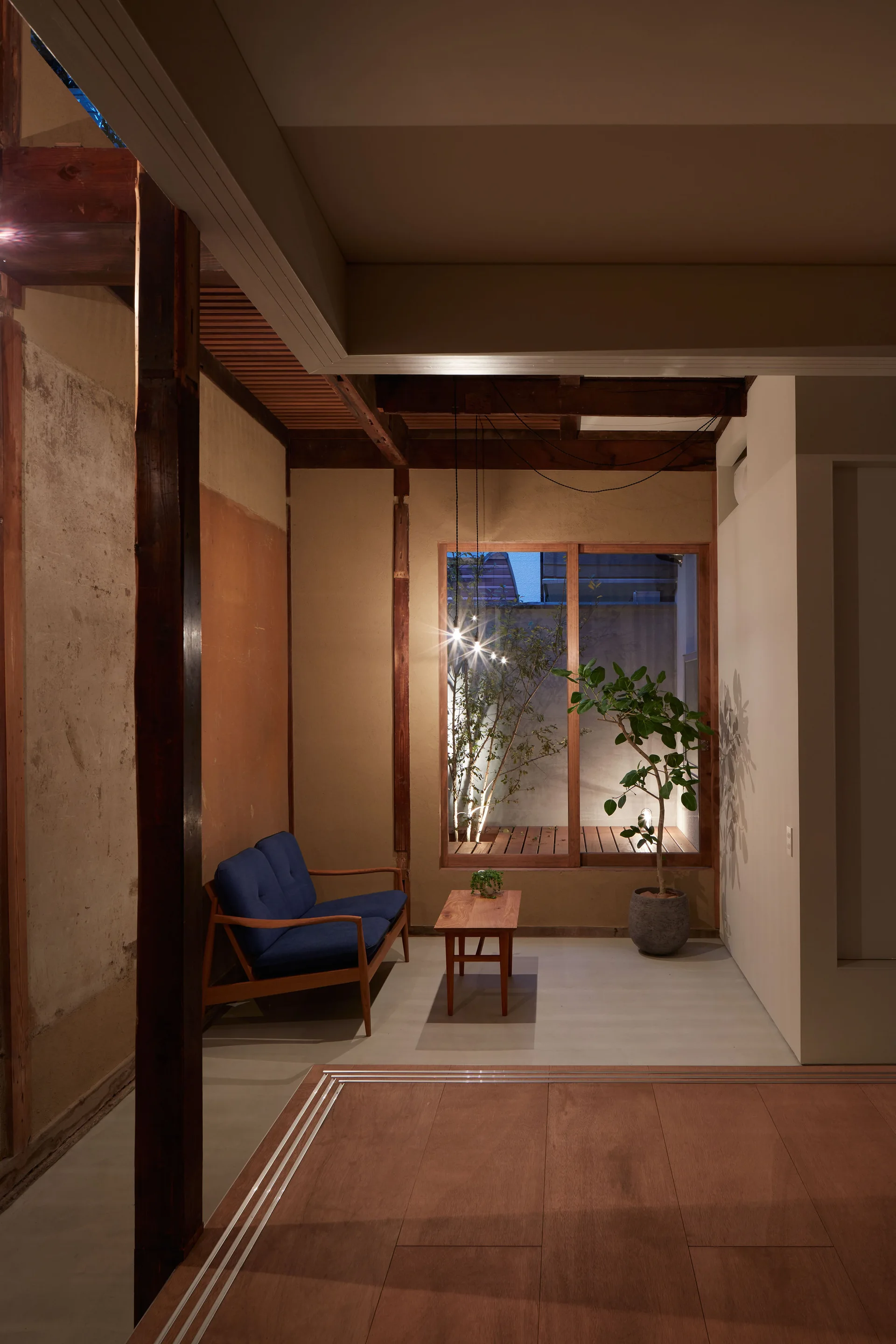 A minimalist living room features a blue wood-frame sofa, a low wooden coffee table, exposed timber posts, and a window looking out to a lit garden.