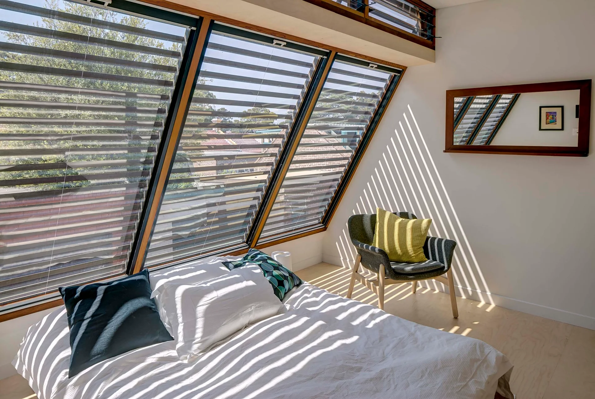 A bright bedroom featuring a sloped window wall with horizontal blinds that cast striped shadows across white bedding and a grey armchair.