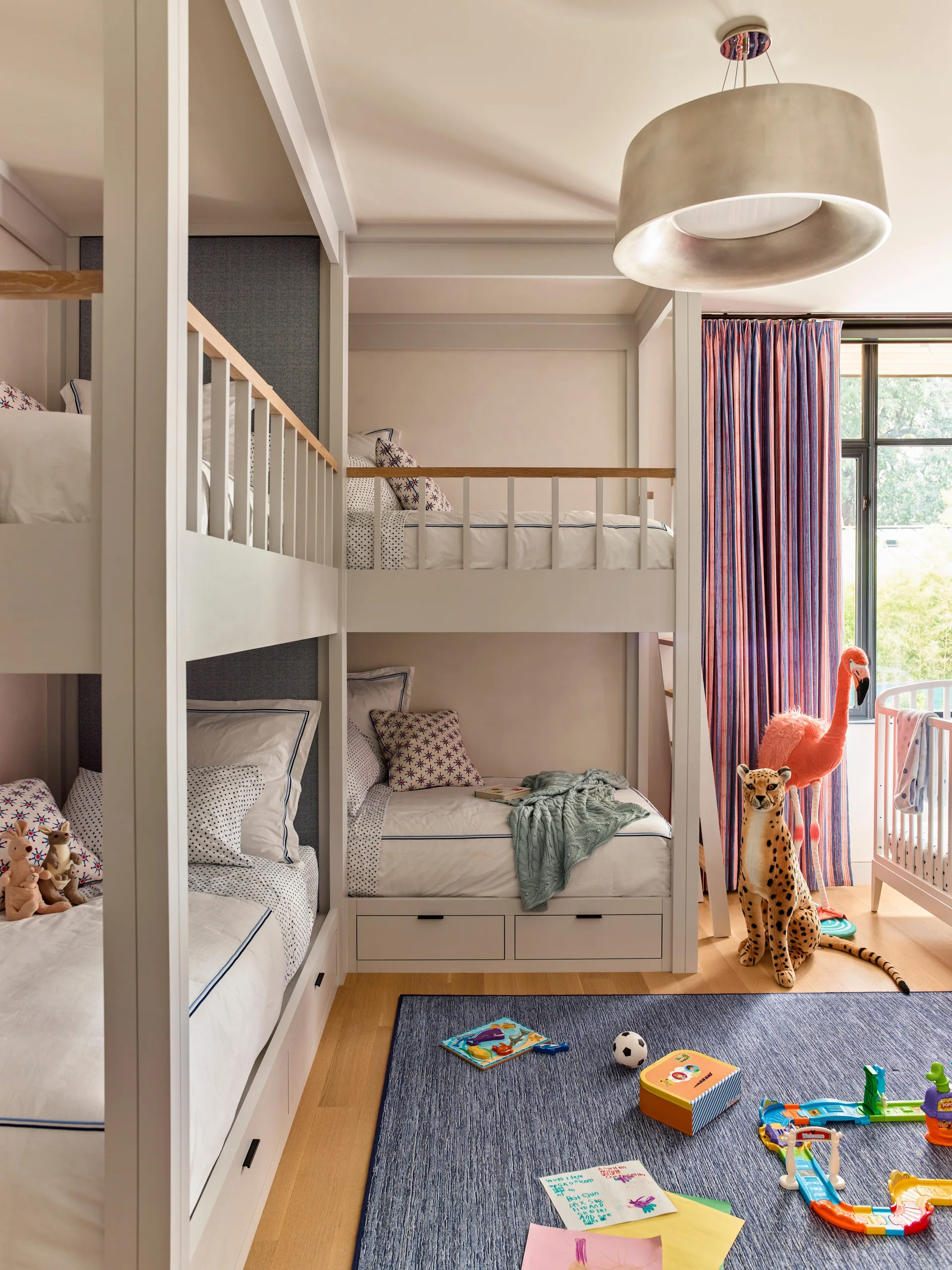 A childrens bedroom features built-in white bunk beds, a blue area rug, colorful toys, striped curtains, and plush animal decor.