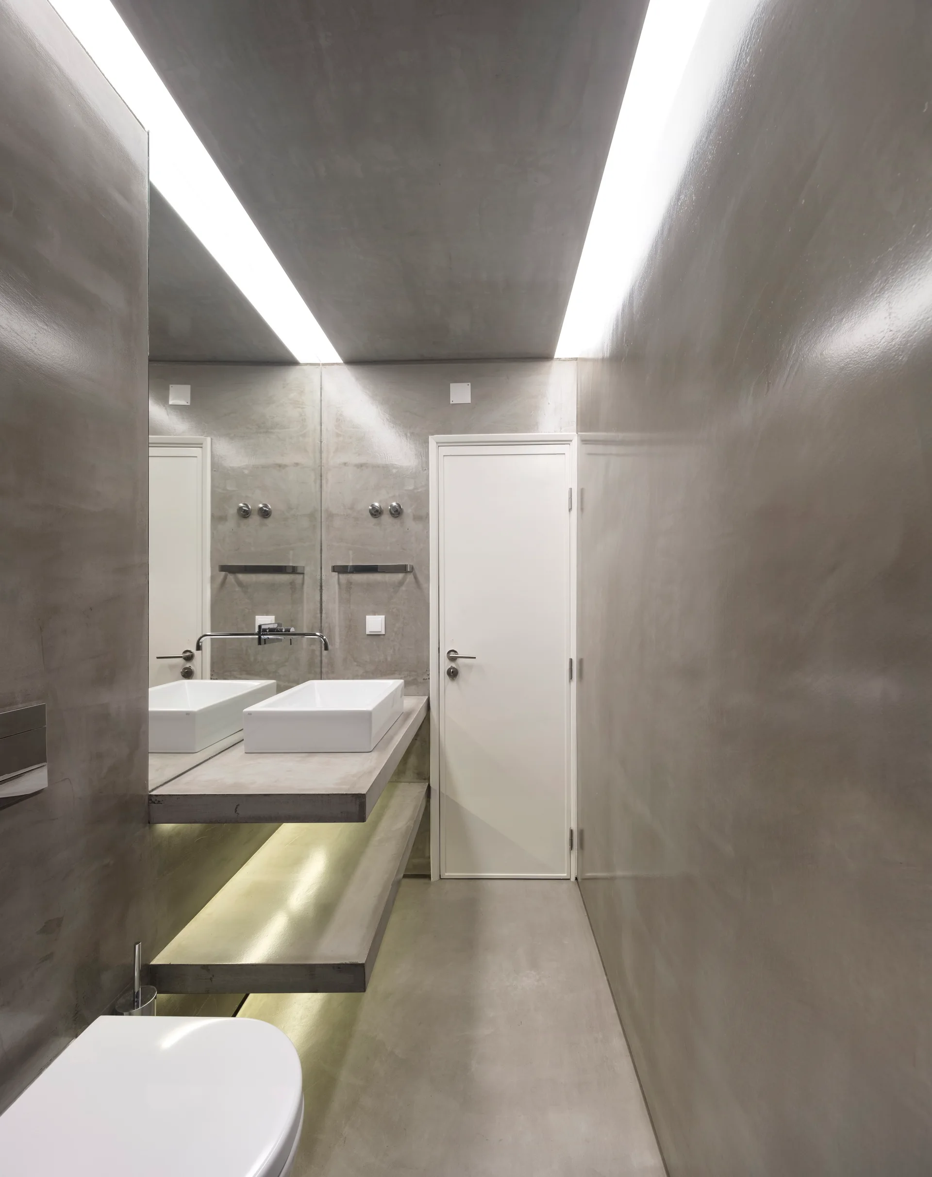 A minimalist bathroom features grey polished concrete walls, a floating concrete vanity with a white vessel sink, and linear cove lighting on the ceiling.