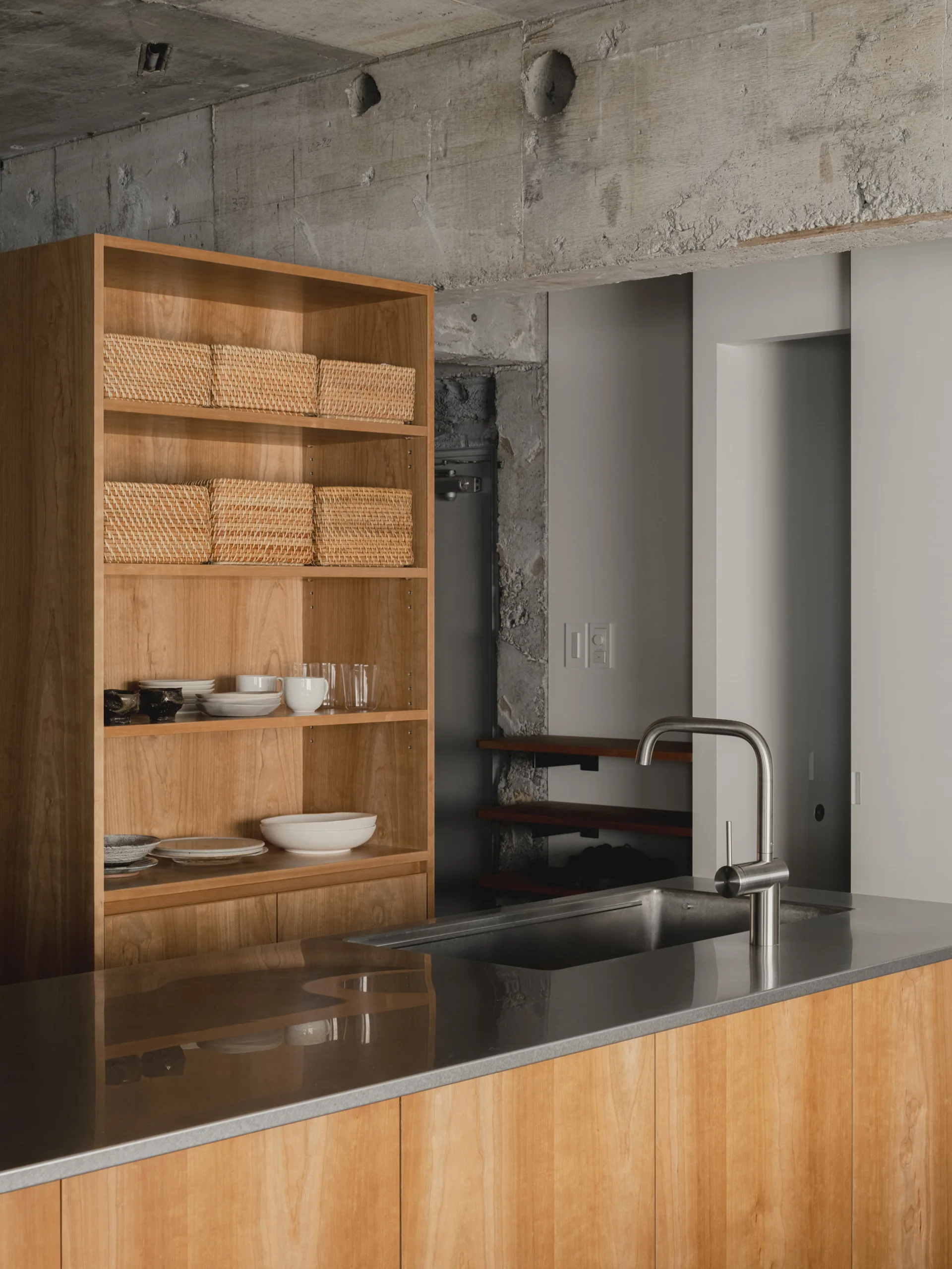 A minimalist kitchen featuring a wood island with a stainless steel sink, set beneath an exposed concrete ceiling and next to a tall wooden shelving unit.