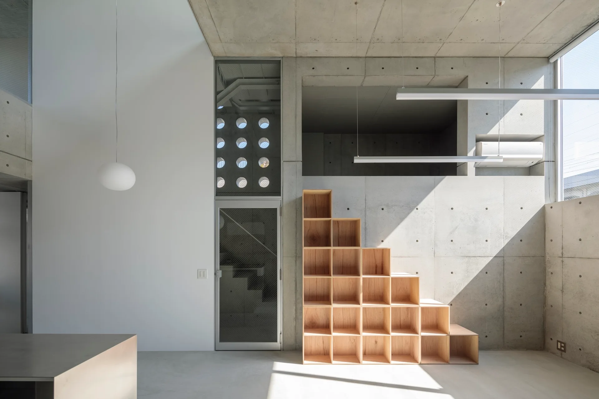 A minimalist concrete interior features a stepped wooden shelving unit, a glass door, and circular cutout windows.