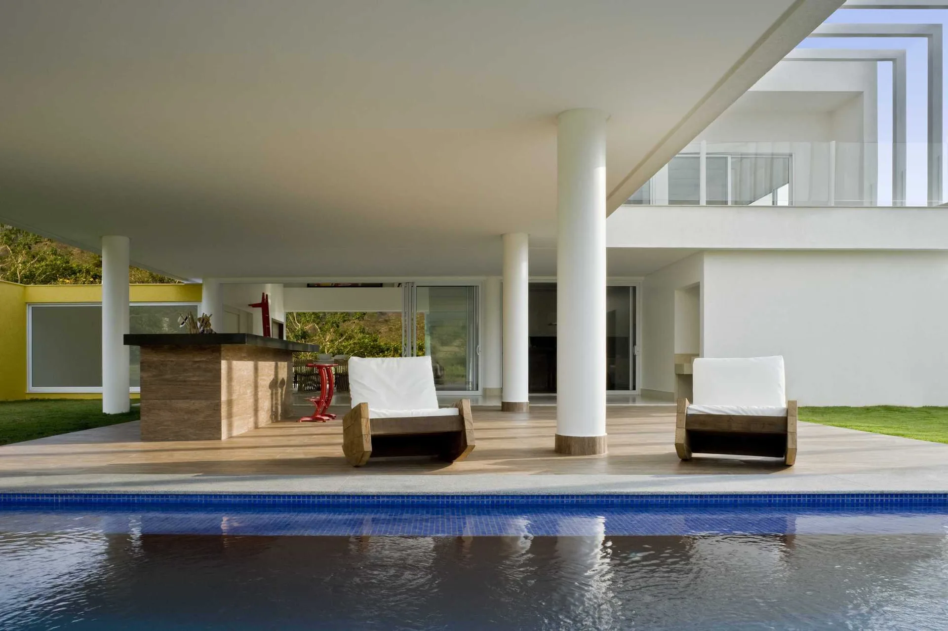 Two modern wooden lounge chairs with white cushions sit on a wood-look deck under a deep white ceiling overhang next to a swimming pool.