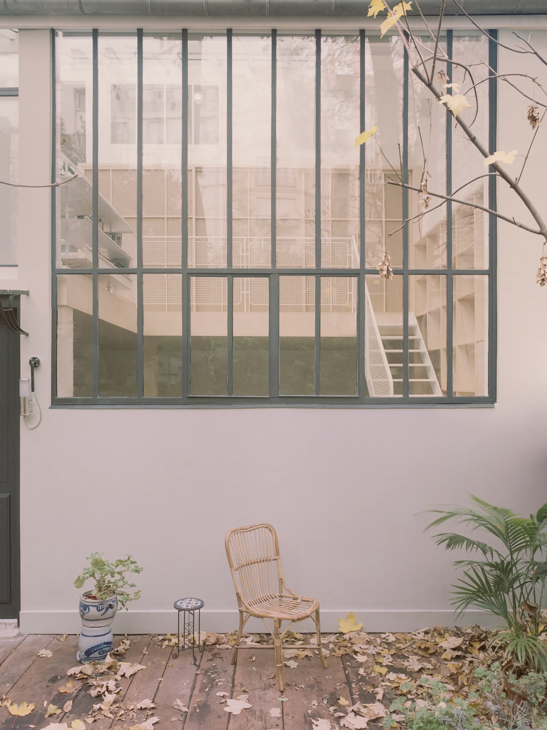 A natural wicker chair sits on a weathered wooden deck next to a white wall with a large steel-grid window.