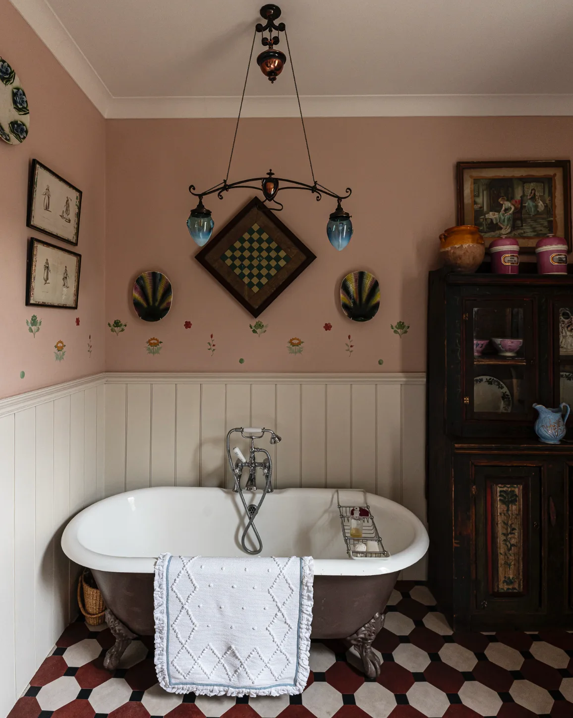 A vintage clawfoot bathtub sits on a geometric tiled floor next to a dark wood cabinet in a bathroom with pink walls and white wainscoting.