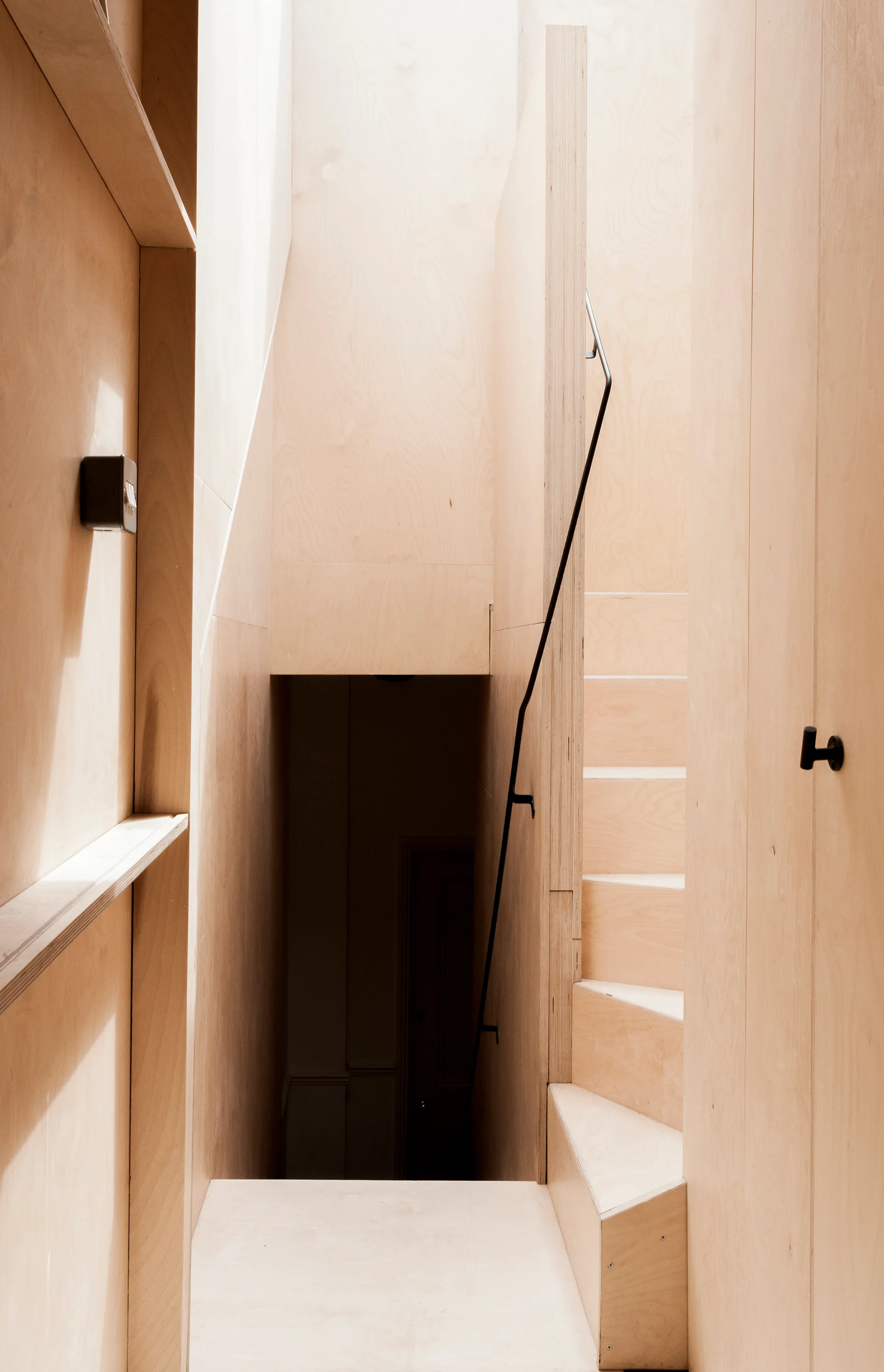 A minimalist stairwell constructed entirely of light beige plywood panels with a thin black metal handrail guiding up the steps.