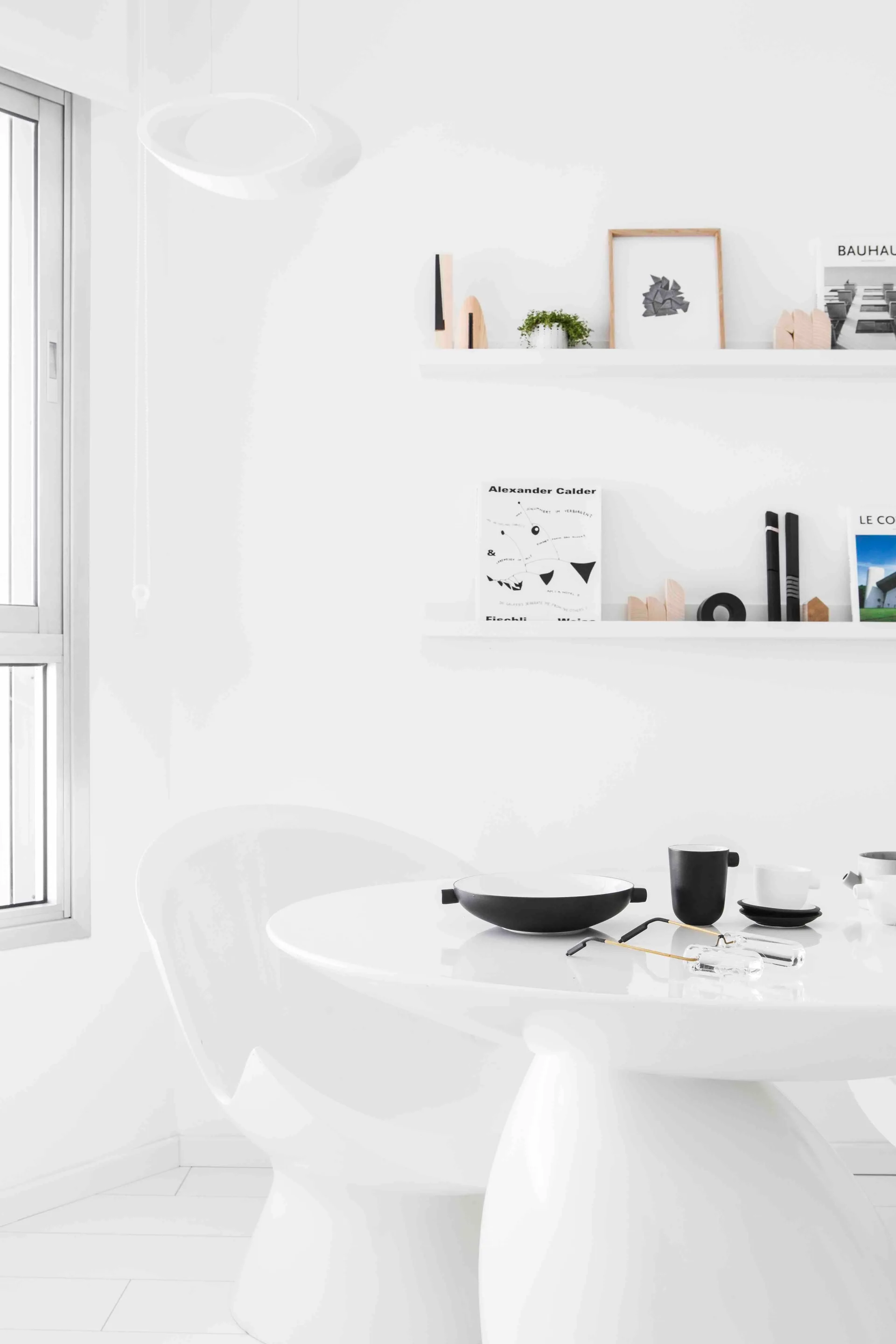 A minimalist white dining room with a glossy pedestal table, a molded plastic chair, and two floating shelves showing books and small decor.