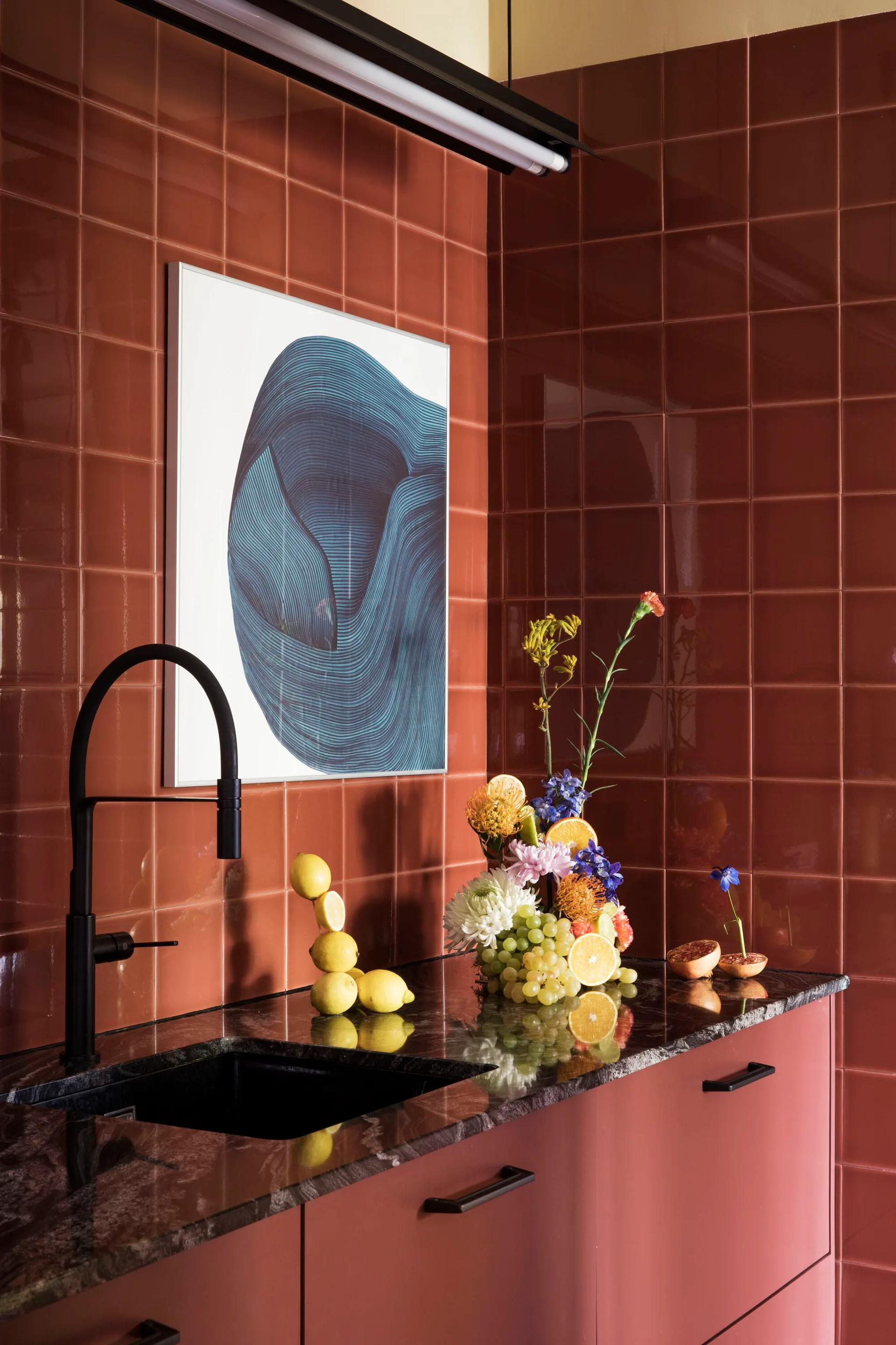 A kitchen corner features a glossy terracotta tiled backsplash, flat-panel cabinets, a black stone countertop with a matte black faucet, and abstract art.