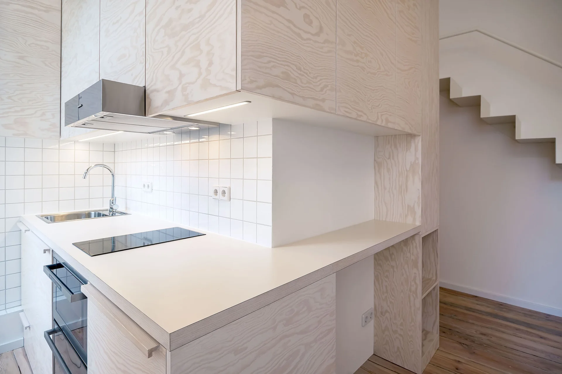 A minimalist kitchen features light-toned plywood cabinets, a white square tile backsplash, an integrated cooktop, and a staircase in the background.