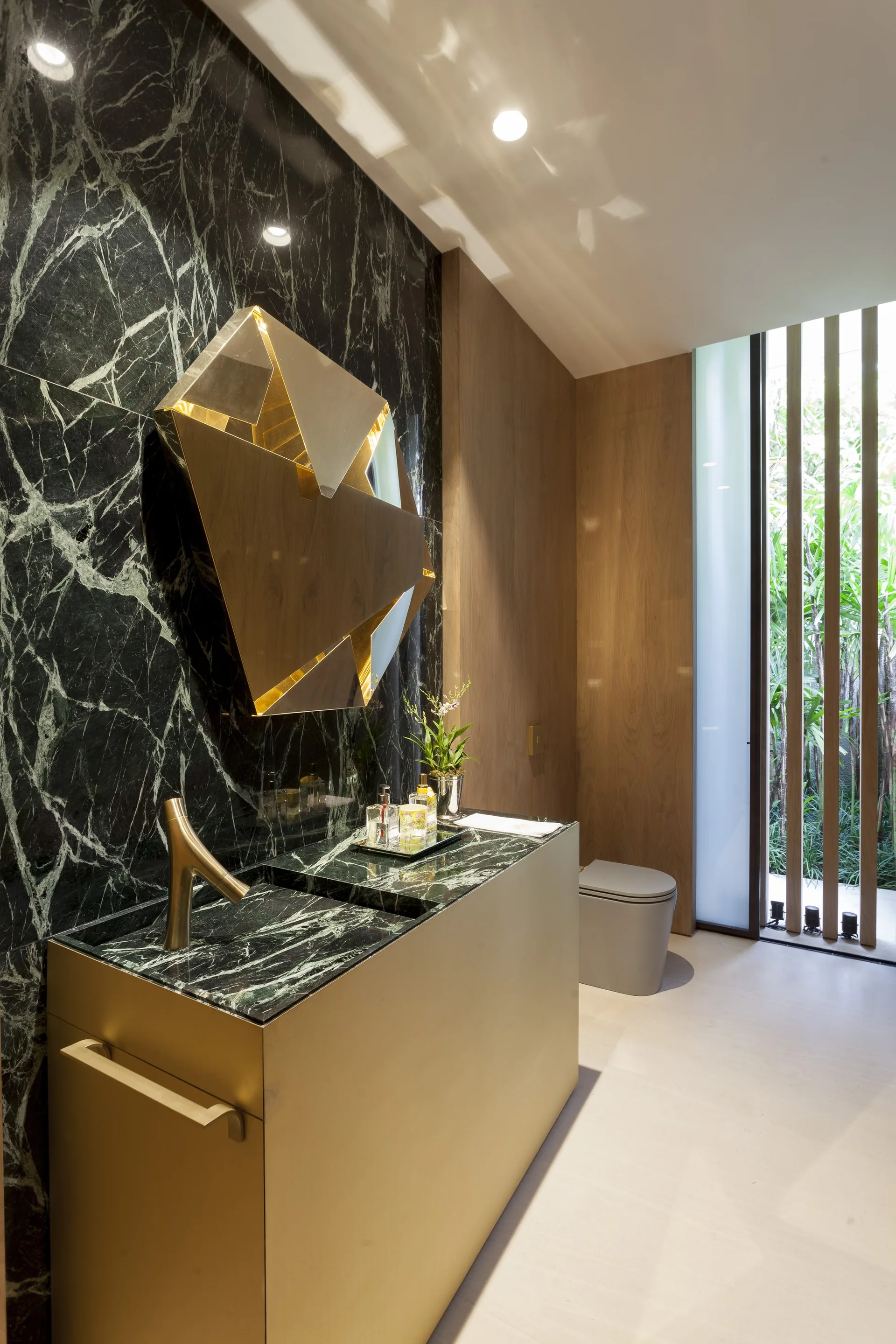 A modern powder room features a gold vanity with a green marble countertop set against a dark green marble accent wall and a faceted gold mirror.