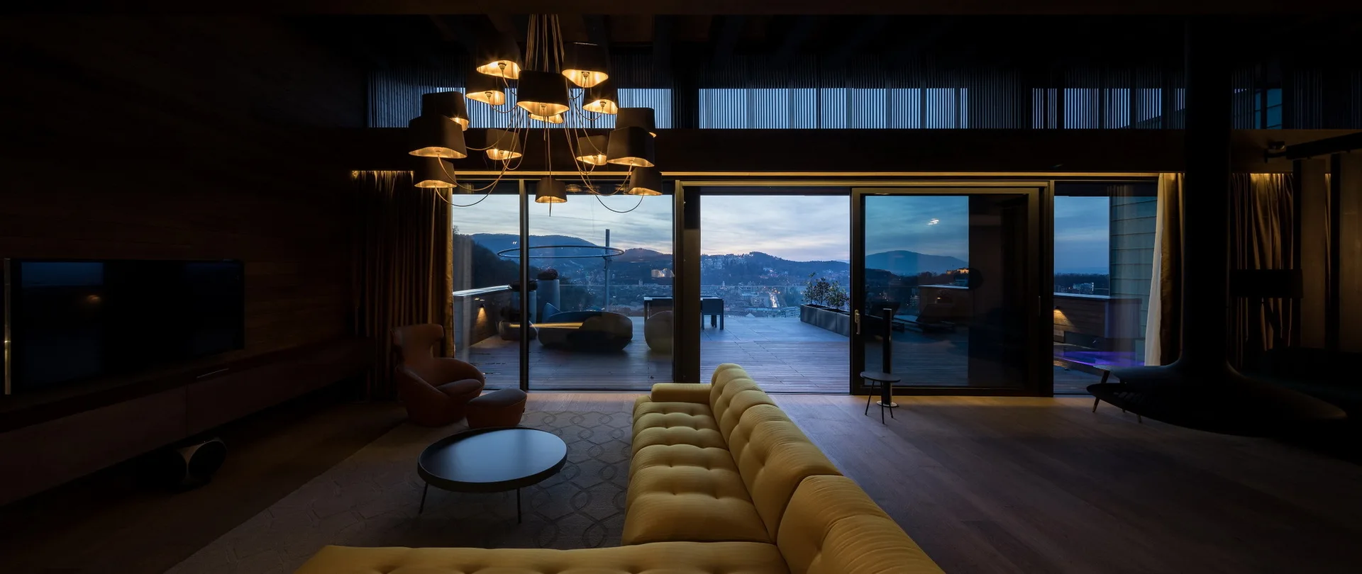 A modern dark wood living room features a bright yellow tufted sectional sofa, a black multi-arm chandelier, and sliding glass doors opening to a terrace.