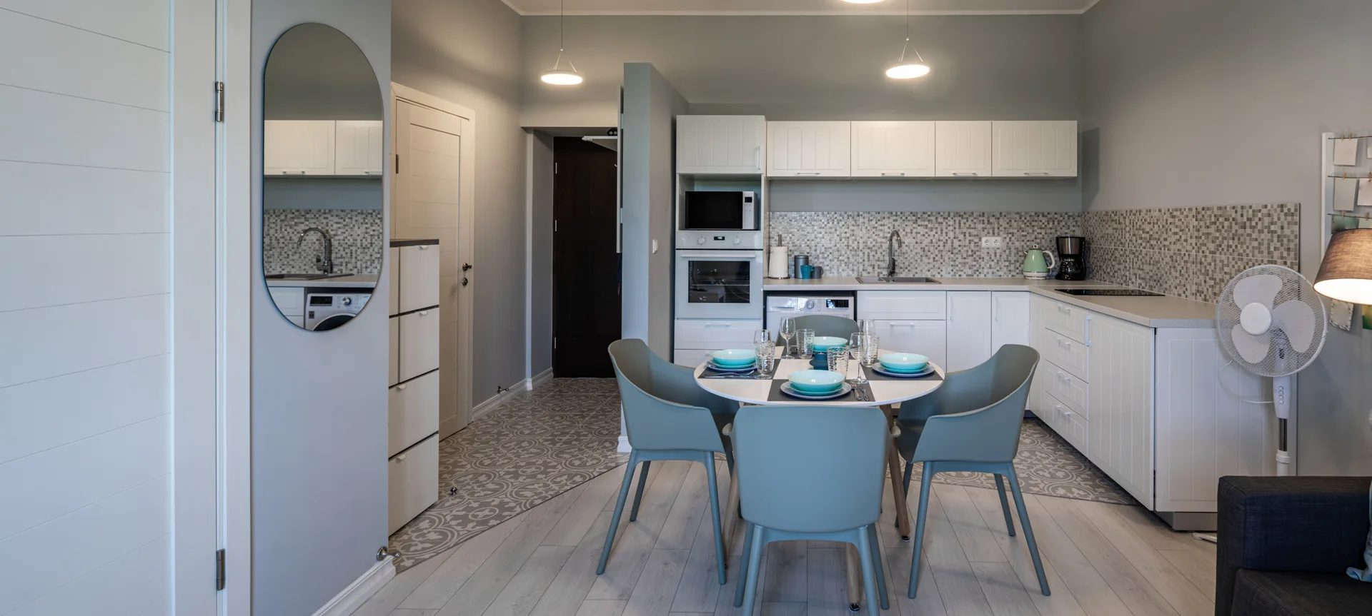 A modern kitchen and dining room featuring a round table with grey chairs, white cabinets, and a diagonal patterned tile floor transition.
