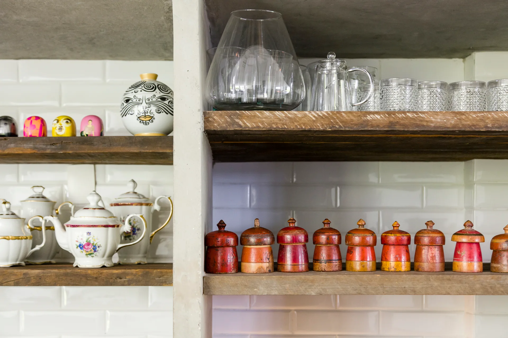 Rustic wood open shelves against a white subway tile backsplash display vintage ceramic teapots, glassware, and small red wooden jars.