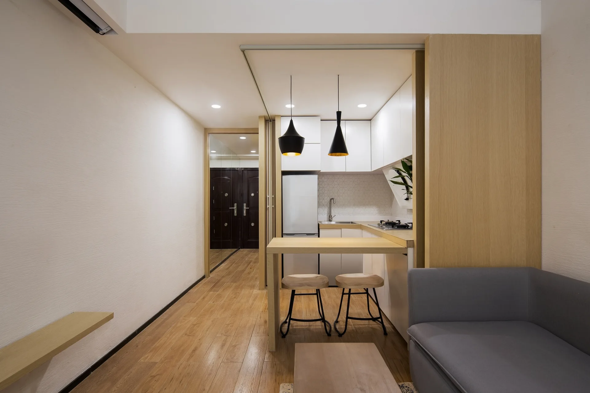 A small kitchen with white cabinets, a light wood peninsula counter, two wooden stools, and black pendant lights next to a grey sofa.