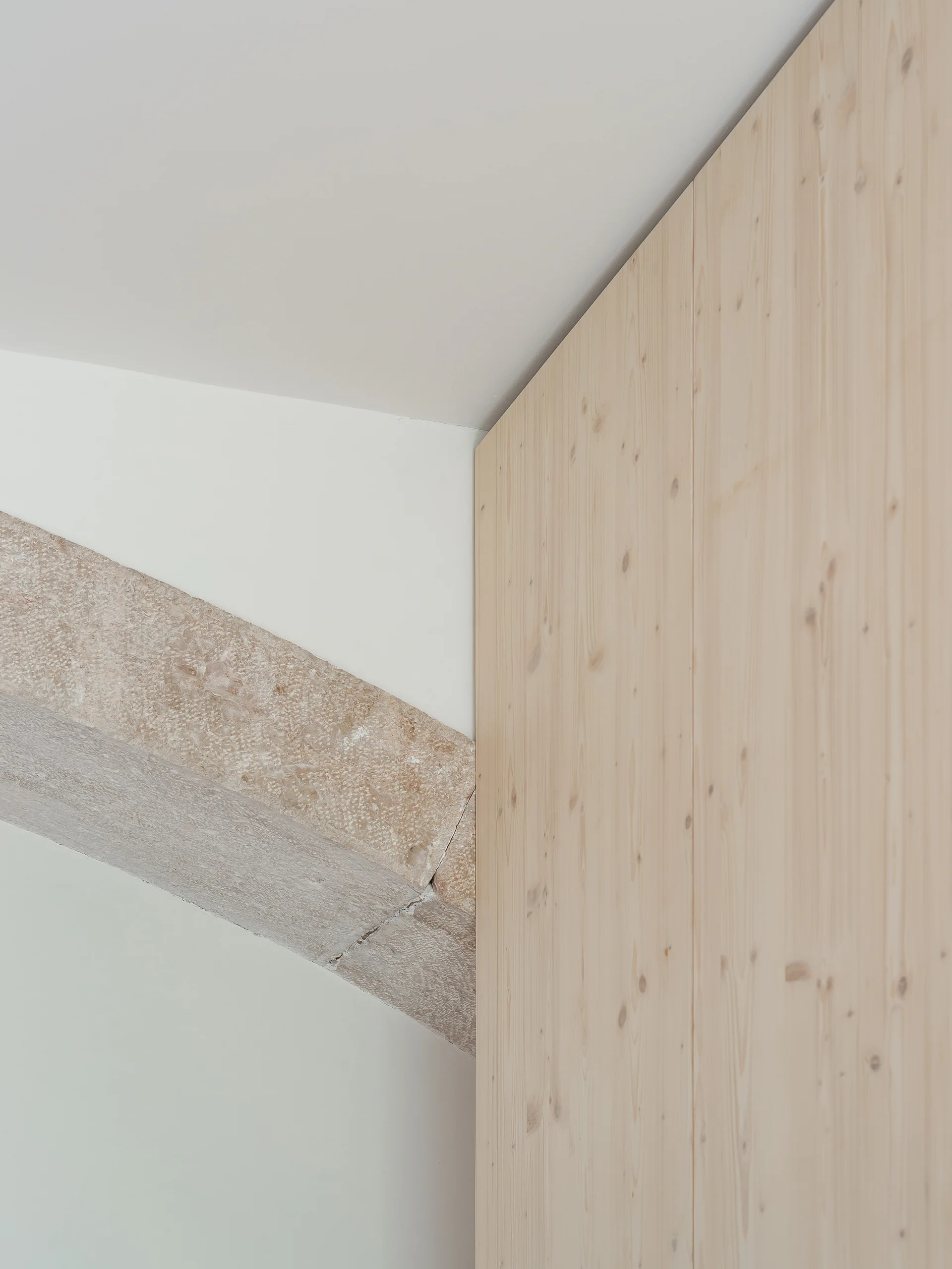 A close-up of light vertical wood cabinetry panels meeting a textured stone beam and a smooth white wall in a minimalist interior.