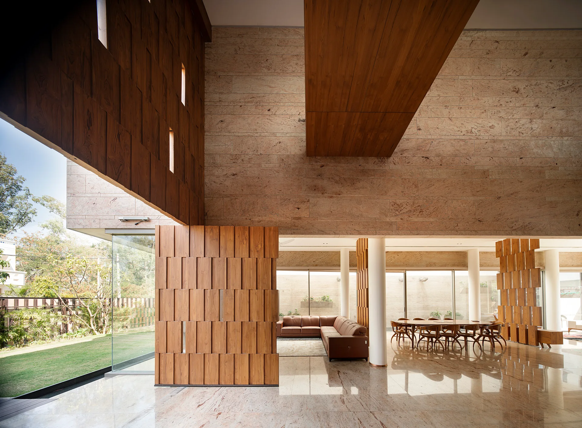 A spacious open-plan interior featuring beige stone-clad walls, polished floors, a brown leather sectional, a dining set, and slatted wooden partitions.