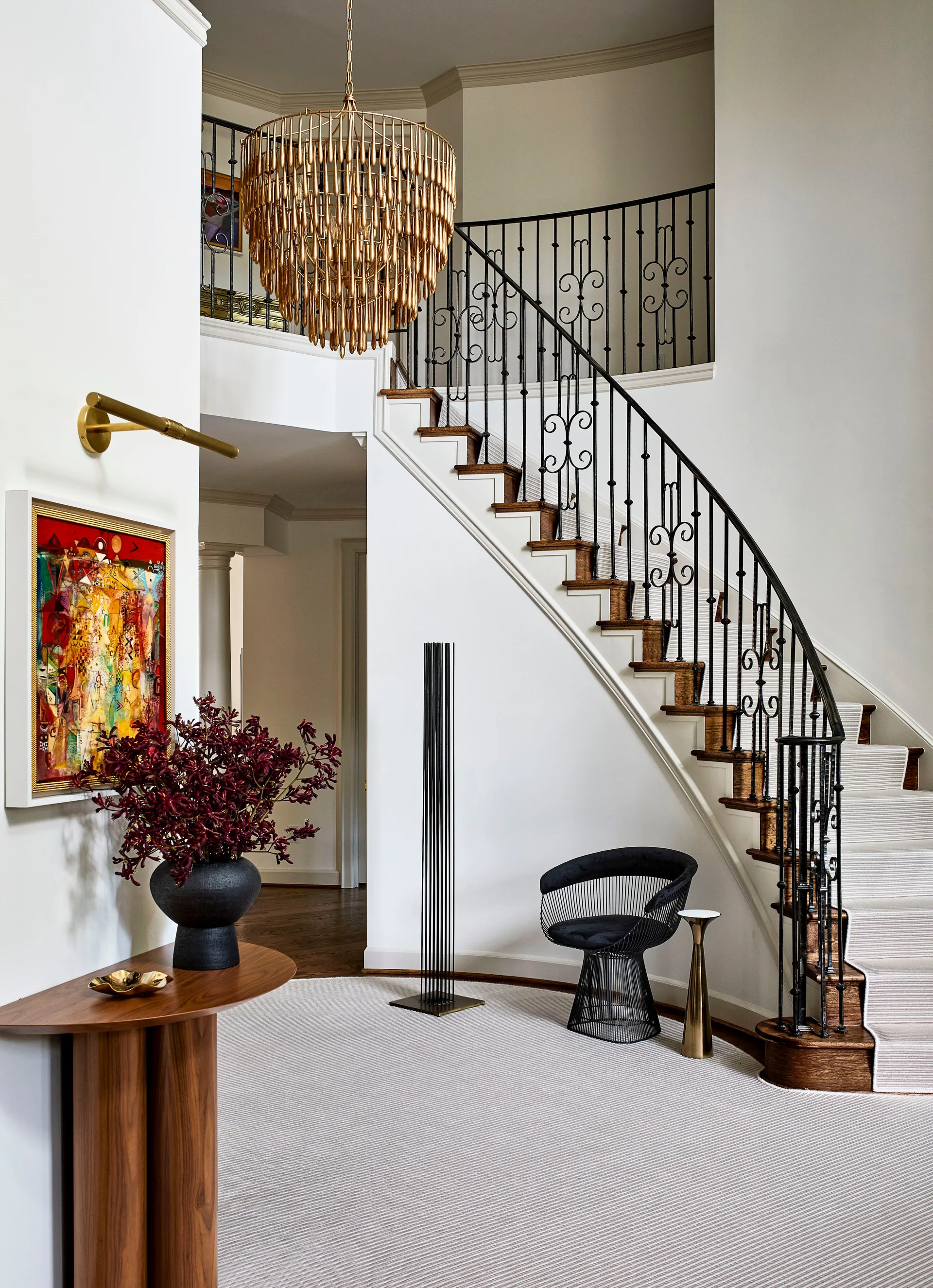 A curved wooden staircase with a striped runner and black iron railing sweeps upward in a white foyer beneath a large brass tiered chandelier.