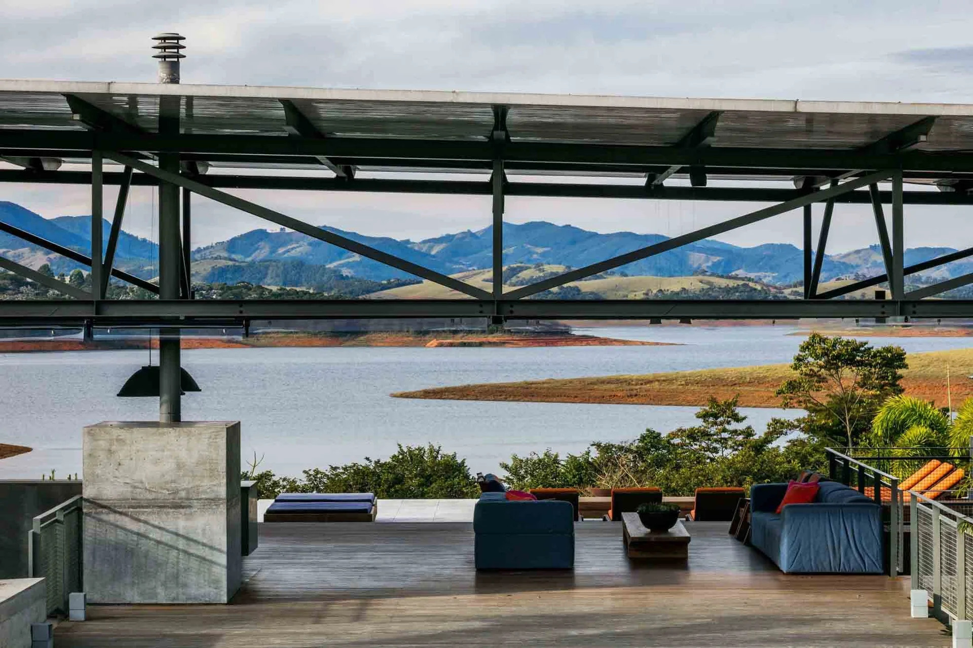 An outdoor lakeside patio featuring blue upholstered sofas on a wood deck under a large industrial steel canopy overlooking a lake and mountains.