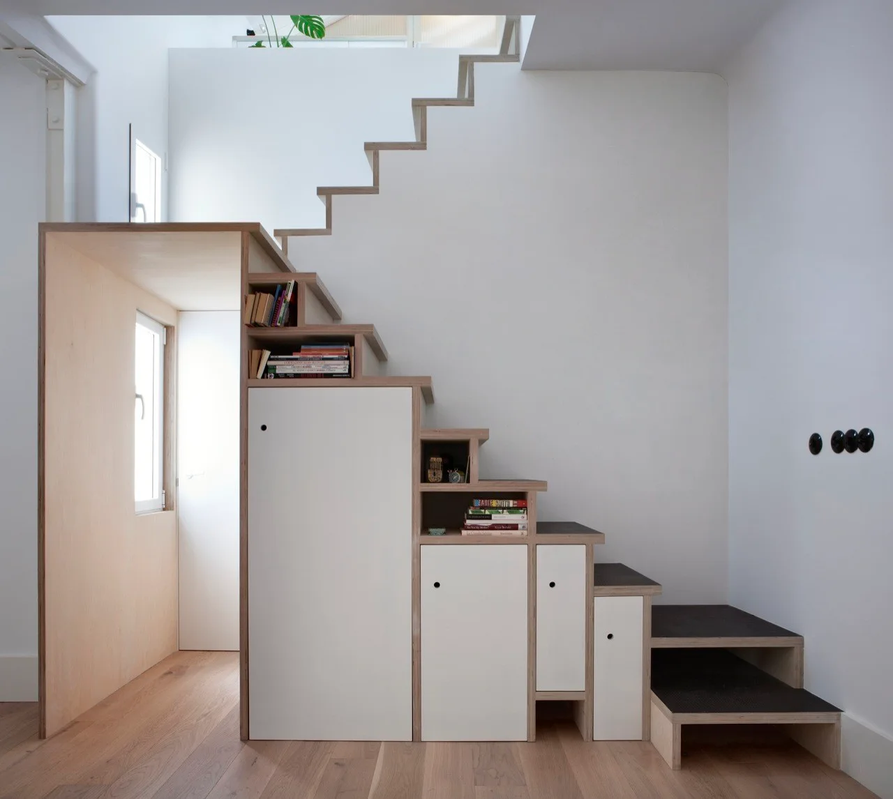 A light wood staircase features integrated white cabinets, small open bookshelves, and dark treads against a plain white wall.