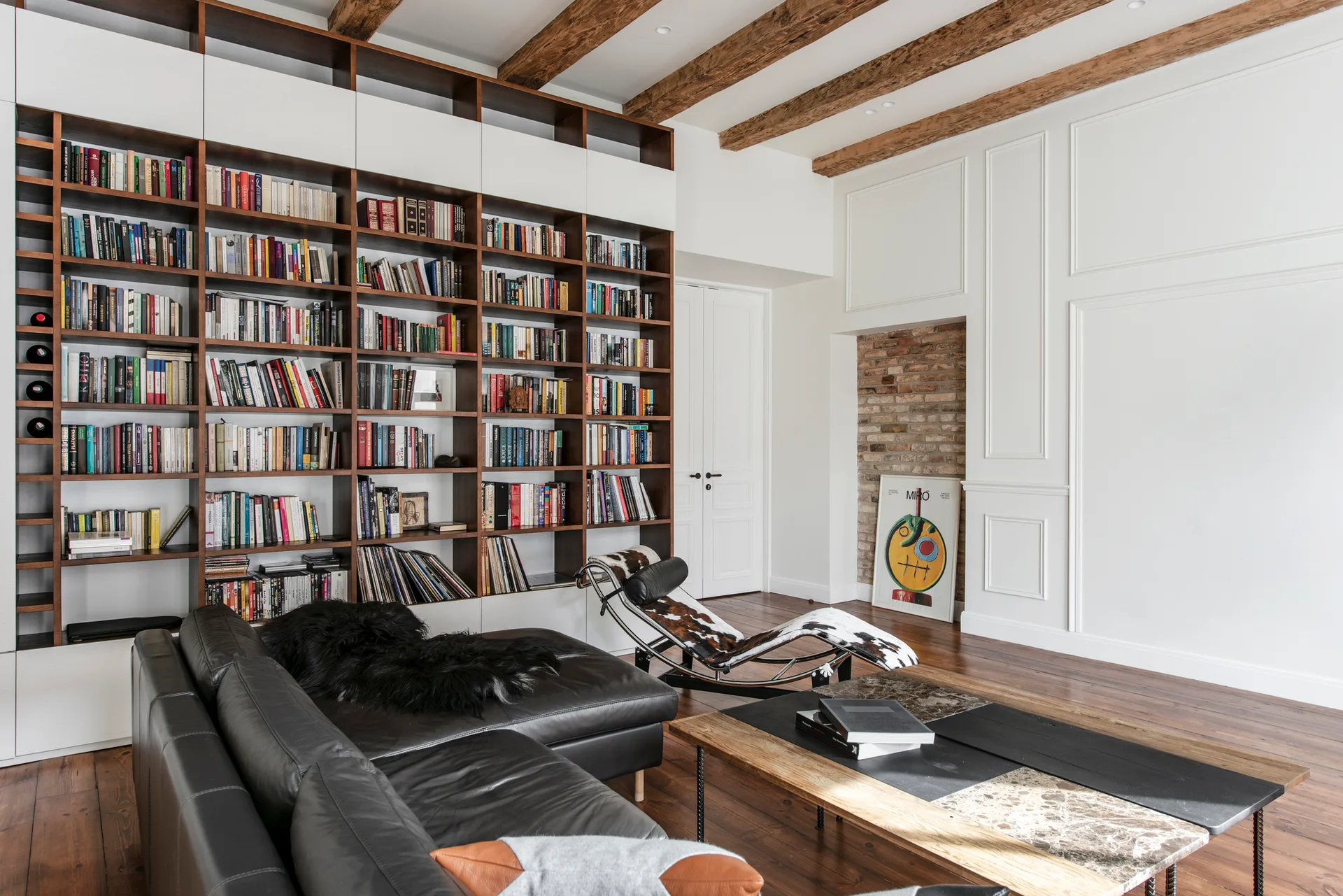 A spacious living room featuring a floor-to-ceiling wooden library bookcase, a black leather sectional sofa, and exposed wooden ceiling beams.