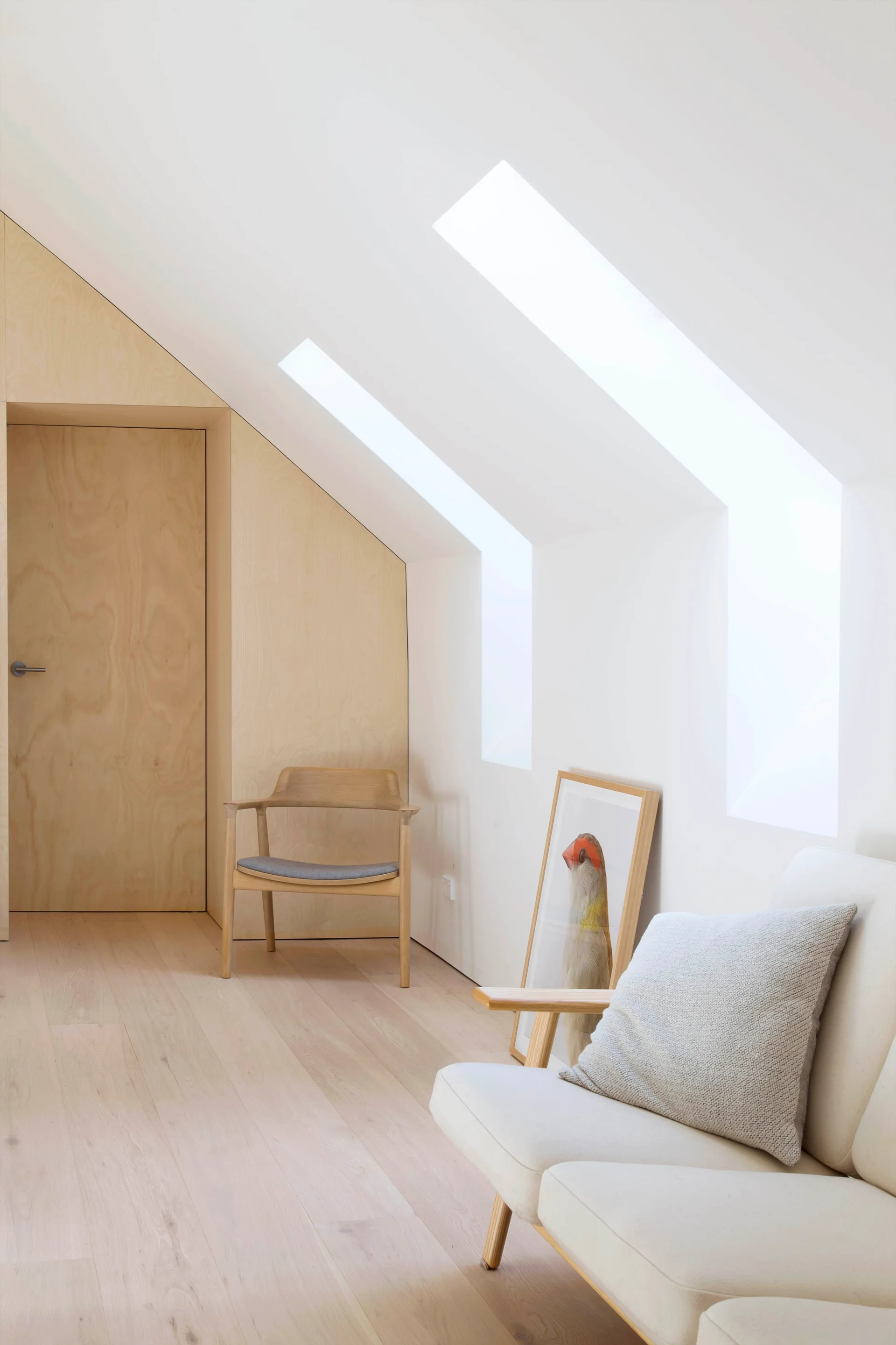 A bright attic sitting area with sloped ceilings, skylights, light wood floors, and a plywood wall with an integrated door.