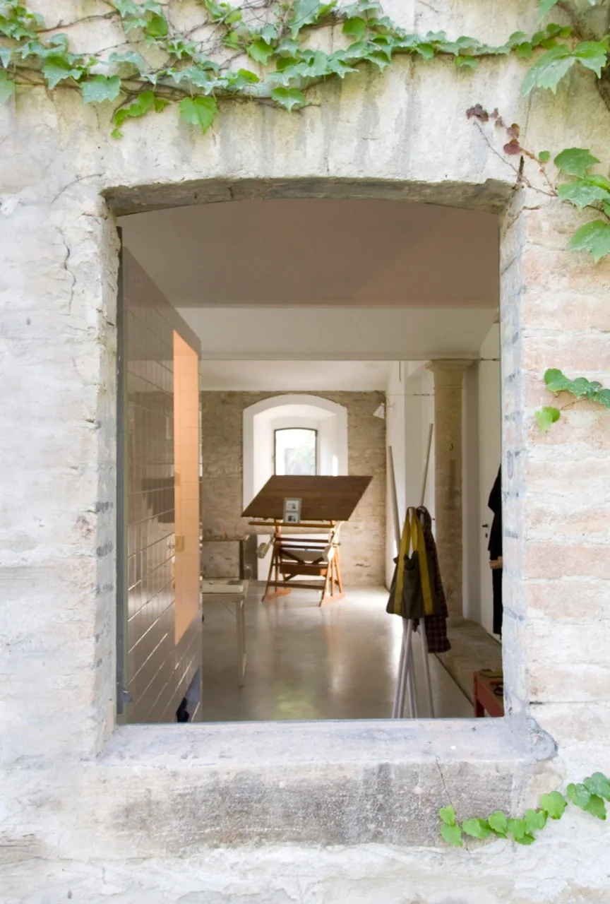 A view through an exterior stone archway looking into a minimalist home studio with a wooden drafting table, concrete floor, and stone walls.