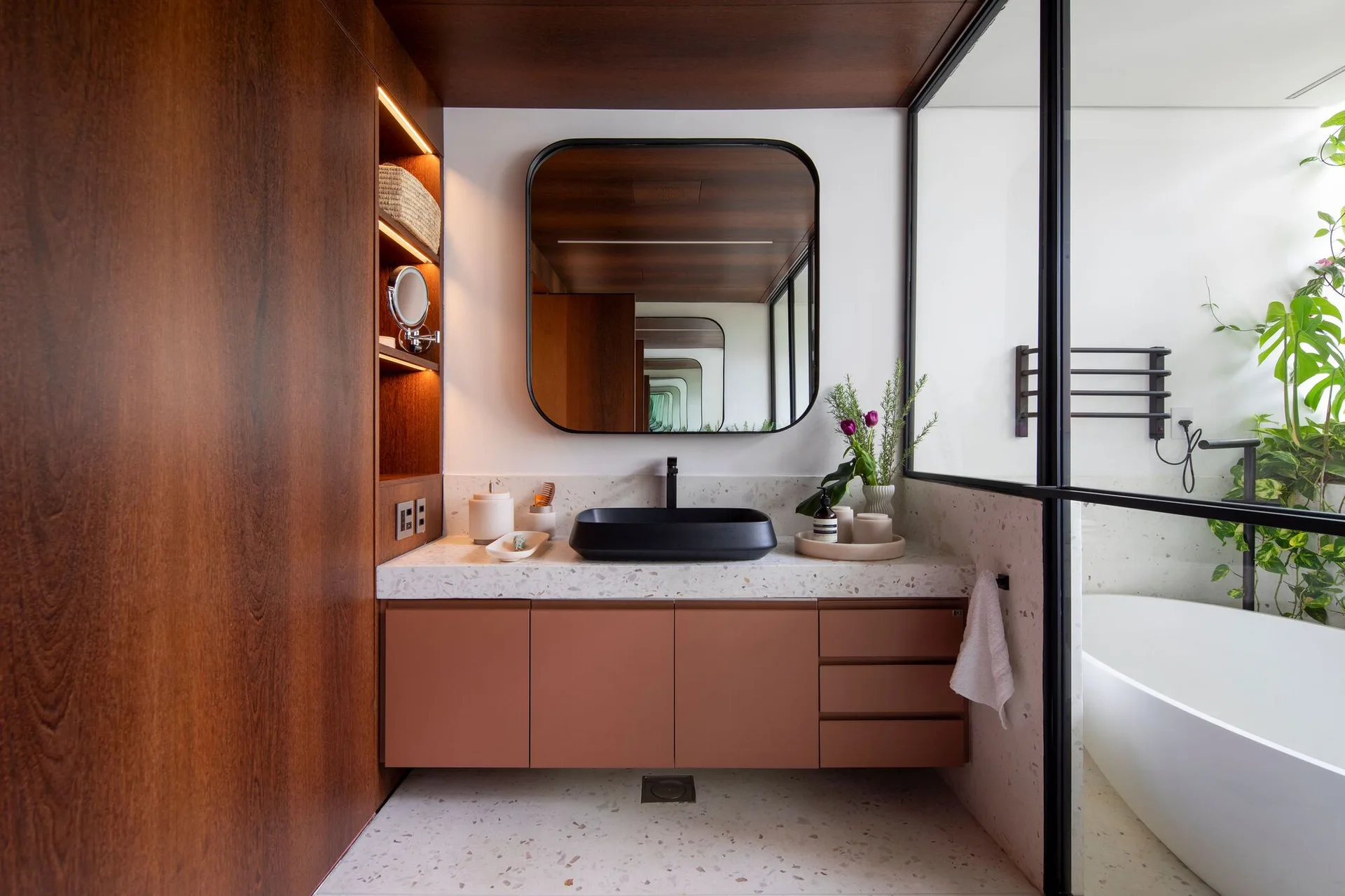 A modern bathroom features a terracotta floating vanity, terrazzo countertop, and black vessel sink, alongside a glass-enclosed bathtub area.