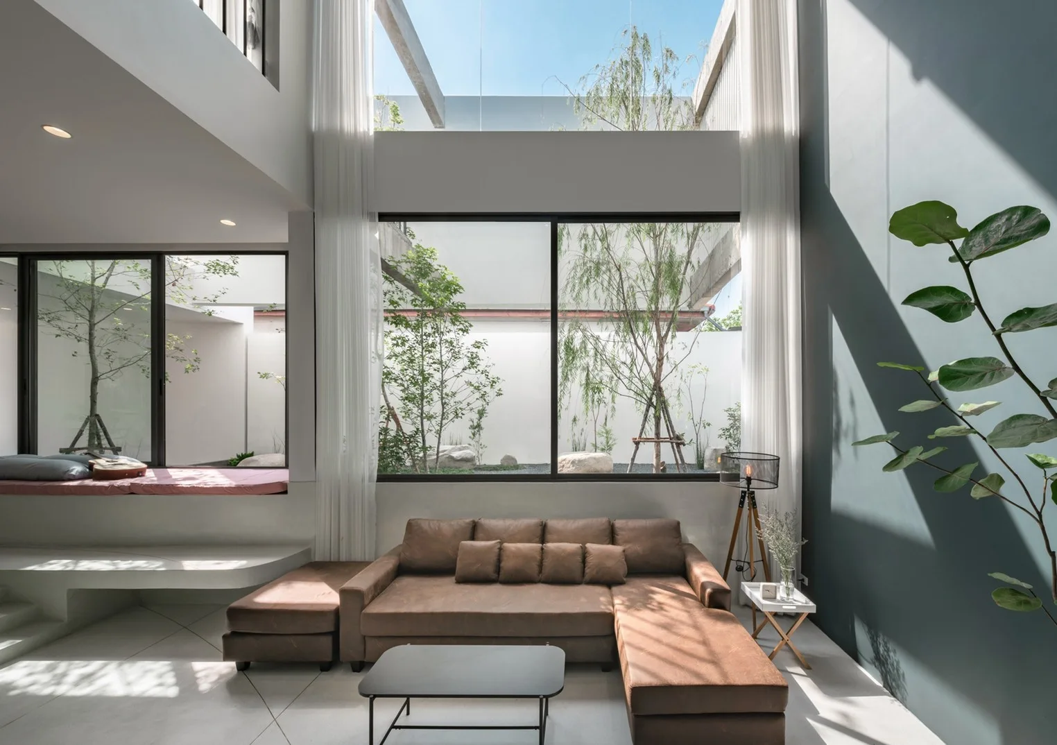 A high-ceiling living room features a brown sectional sofa facing a large window that overlooks an outdoor courtyard with green trees.
