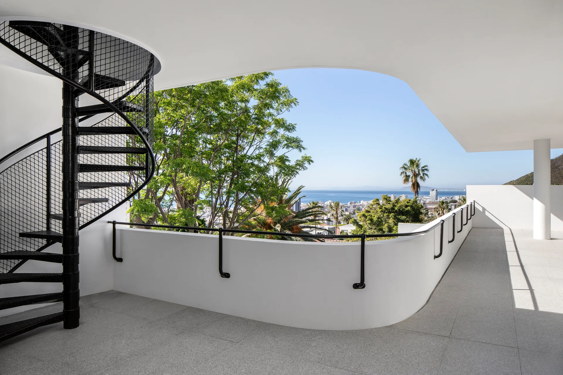 A modern outdoor terrace with a black metal spiral staircase, a white curved balcony wall, and a view of trees and the sea.