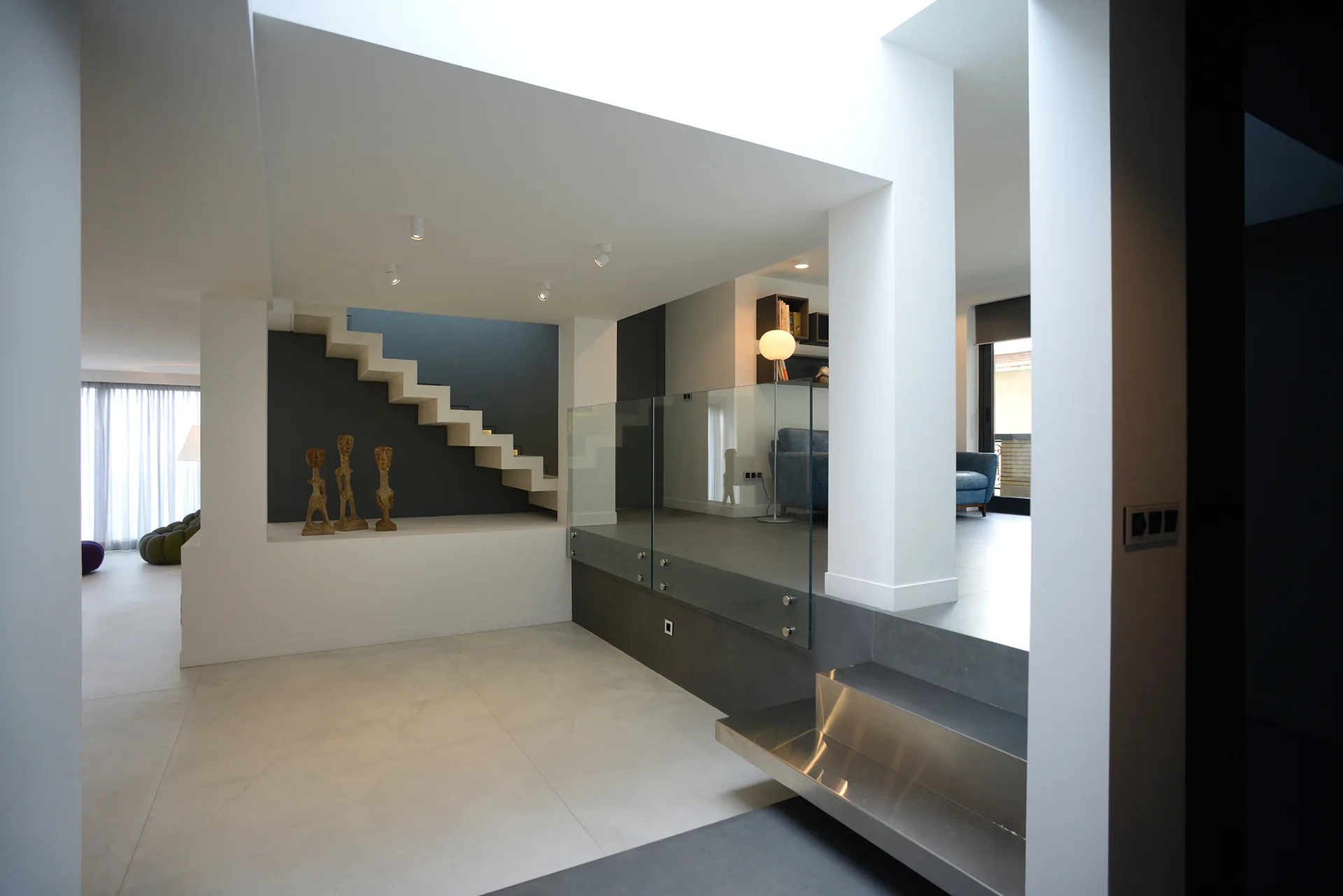 A minimalist split-level interior featuring white floating stairs against a grey wall, a glass railing, and large-format floor tiles.