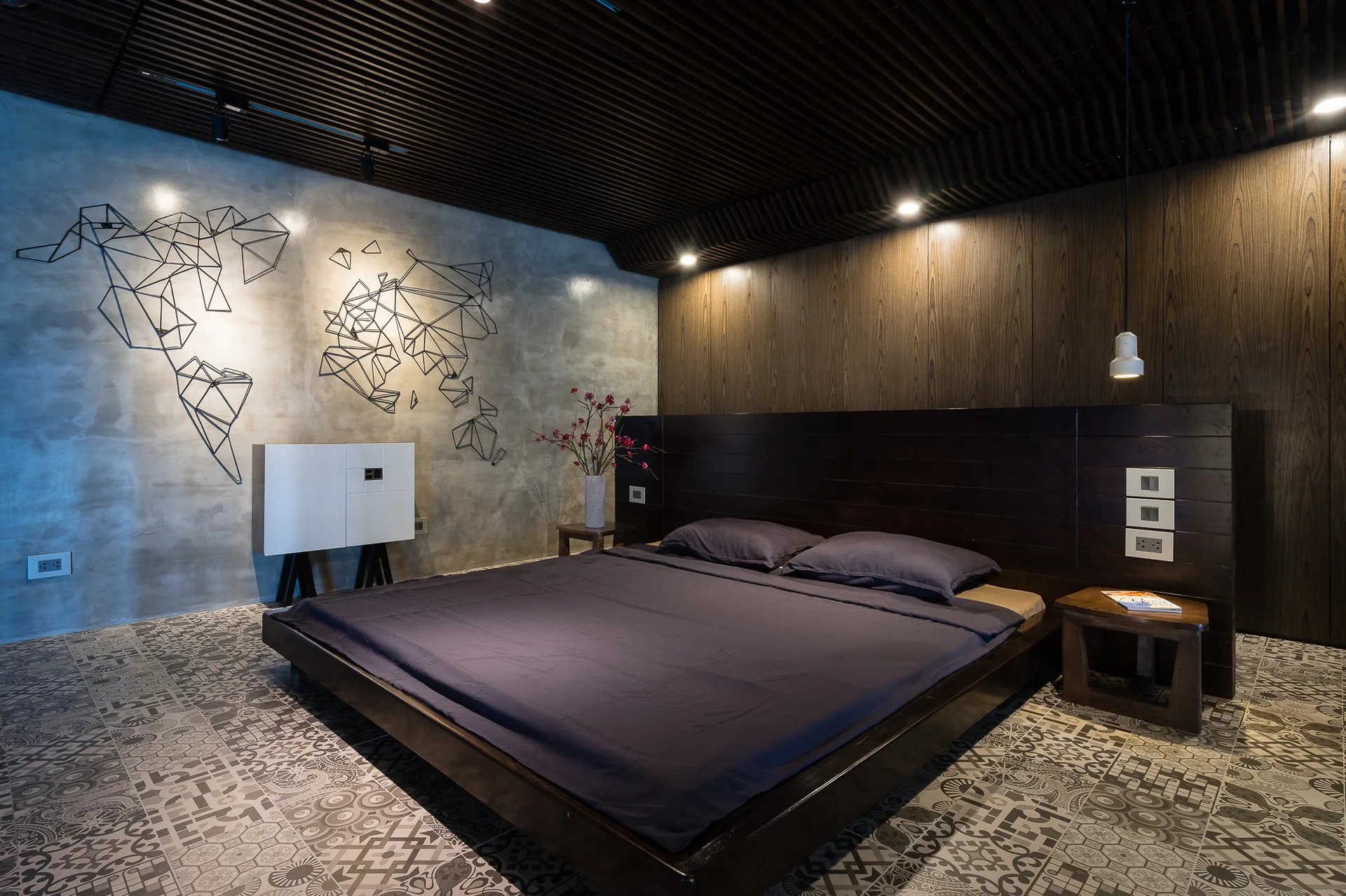 A modern bedroom featuring a dark wood platform bed, a polished concrete wall with geometric art, and patterned ceramic floor tiles.